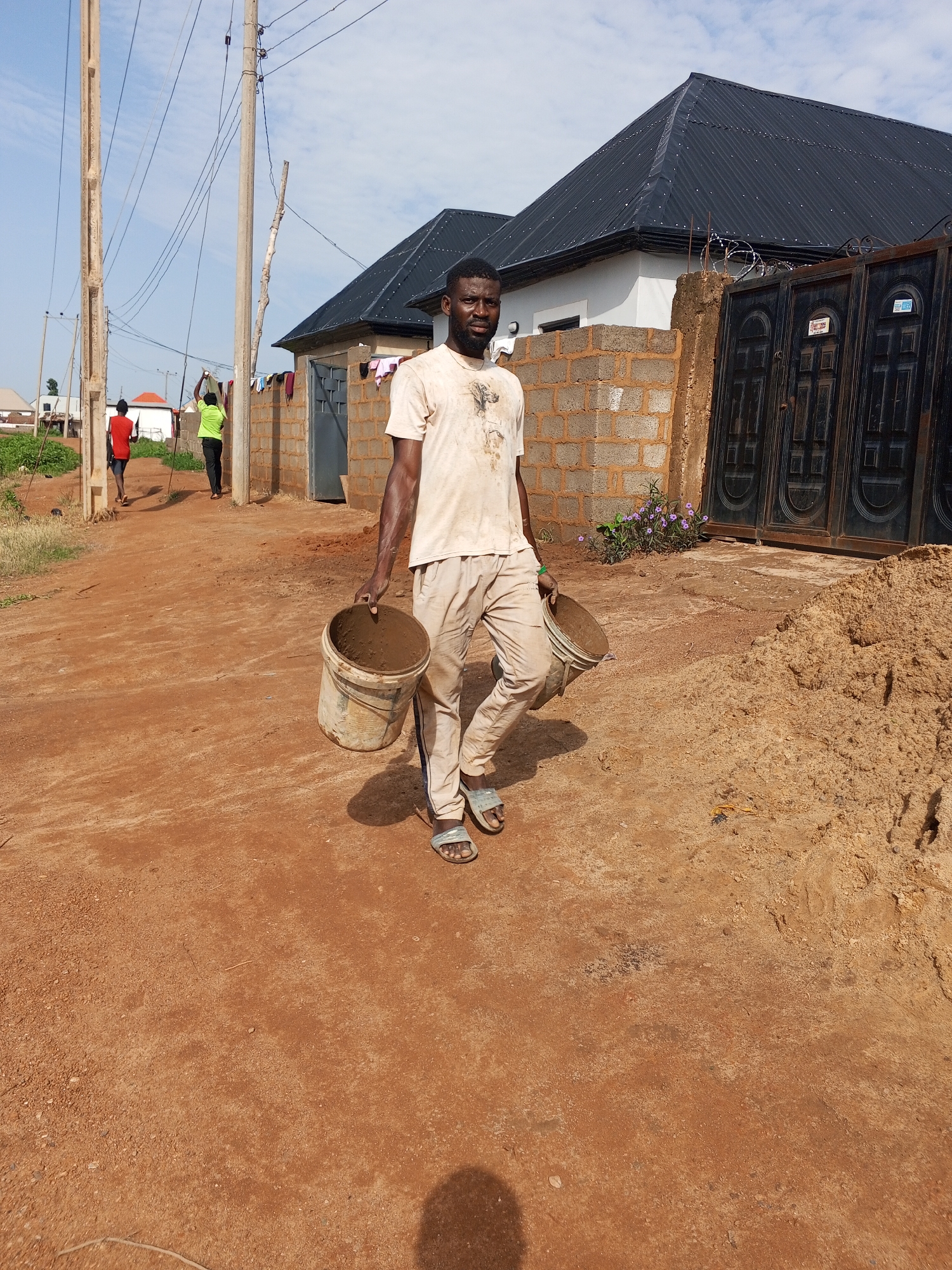 The image depicts a man carrying two buckets of dirt, likely for construction. He is walking on a dirt road in an outdoor environment. In the background, there are buildings with dark roofs, a brick wall, and a metal gate. There are also utility poles with wires. Two other individuals are visible further down the road, one wearing a red shirt and the other a green shirt, also appearing to be walking. The overall scene suggests a construction site or a developing area. The presence of red dirt and the architectural style of the buildings are consistent with certain regions in Abuja, Nigeria.