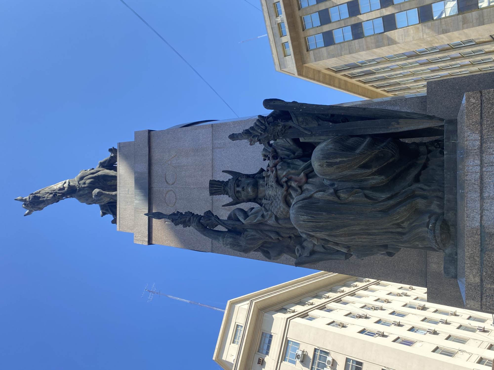 The image shows a low-angle view of the Monument to the Carta Magna and Four Regions of Argentina, also known as the Monument of the Spaniards, located in the city of Buenos Aires, Argentina. The monument, dedicated to the Spanish community on the centenary of the May Revolution, is a tall structure with bronze figures. A bronze statue of a horse and rider is on the top, with additional bronze figures sculpted on the lower sections. The monument is set against a clear blue sky, framed by buildings on either side with numerous windows. A white contrail streaks the sky.
