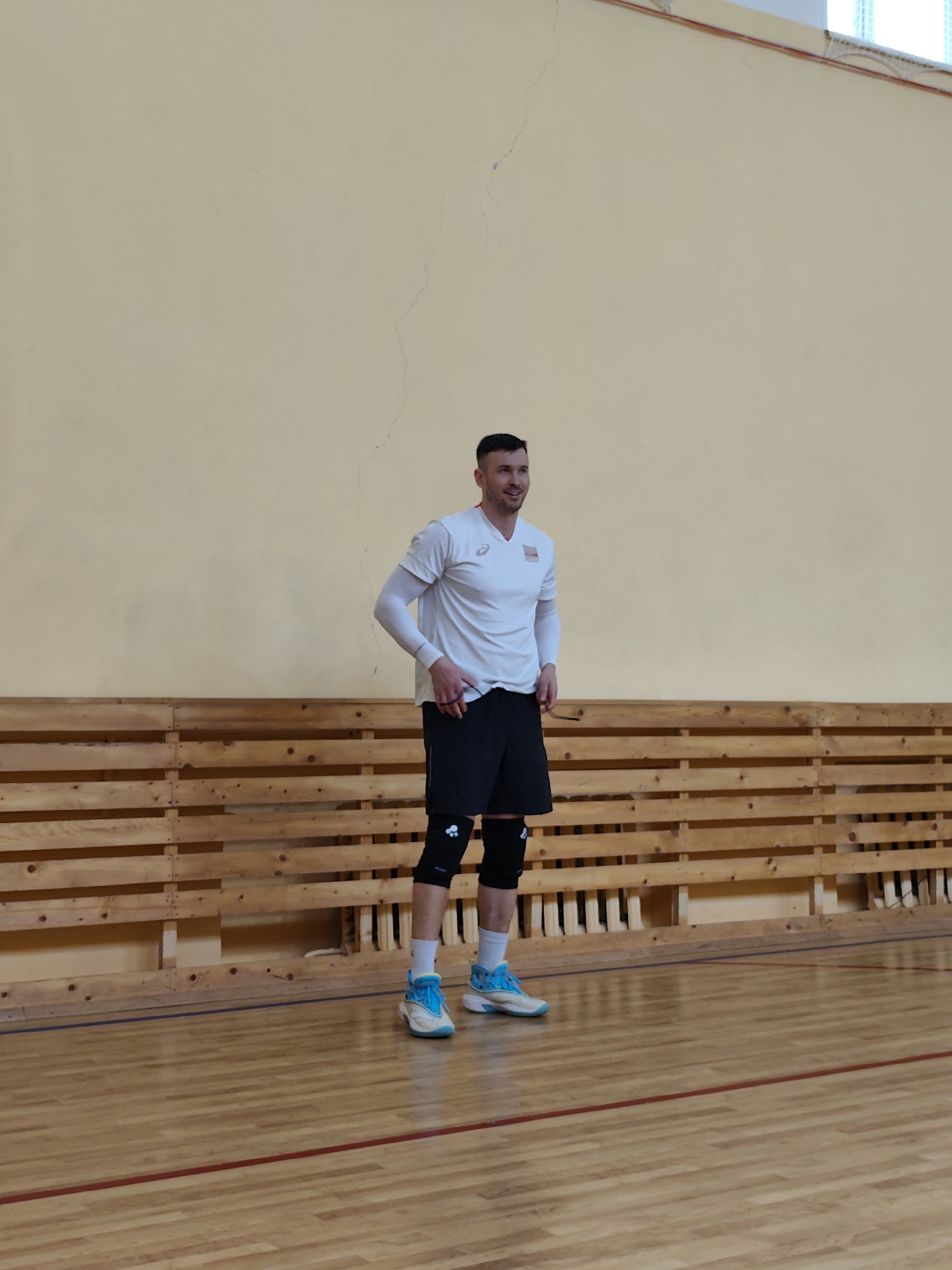 The image captures a male athlete standing in an indoor gymnasium. The subject is positioned slightly to the left of the frame, facing forward but looking towards the right with a pleasant, slight smile. He has short, dark hair. He is dressed in athletic wear, including a white short-sleeved t-shirt with a visible "ASICS" logo and a small emblem on the left chest, white long-sleeved undershirts, black athletic shorts, black knee pads adorned with a white symbol, white socks, and distinctive light blue and white athletic shoes. His hands are resting casually on his hips or waist.
The setting is an indoor sports facility, likely a volleyball or basketball court, indicated by the polished wooden floor with clearly visible blue and red boundary lines. The background consists of a plain light yellow or beige wall, below which runs a horizontal structure of natural wood slats. The lighting appears to be typical for an indoor sports arena. The man's attire, particularly the knee pads, suggests involvement in a sport such as volleyball. The relaxed pose and smile imply he is either resting, observing, or enjoying the activity. The location context is an Unknown, Unknown indoor sports venue.