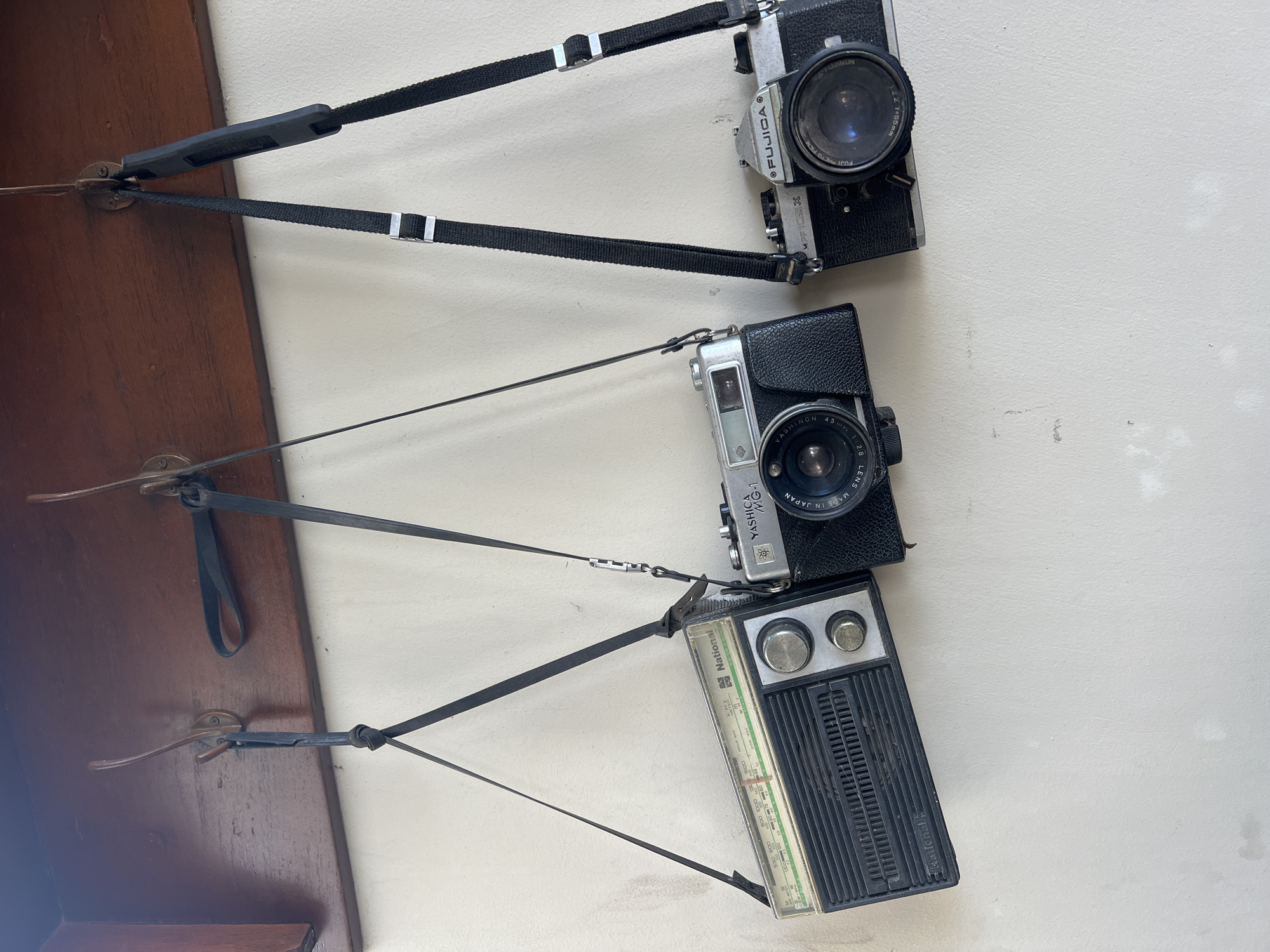 The image shows three vintage items suspended from a wooden board against a white wall, likely within a domestic setting in Ubud District, Indonesia. From top to bottom, the items are:
1. A black Fujica camera with a strap.
2. A silver Yashica MG-1 camera with a black leather covering and a strap.
3. A National radio with tuning dials and a strap.
The objects are connected via straps to hooks on the wooden board. The wooden board appears to be a shelf or part of a display. The overall impression is of a display or collection of vintage electronic items.