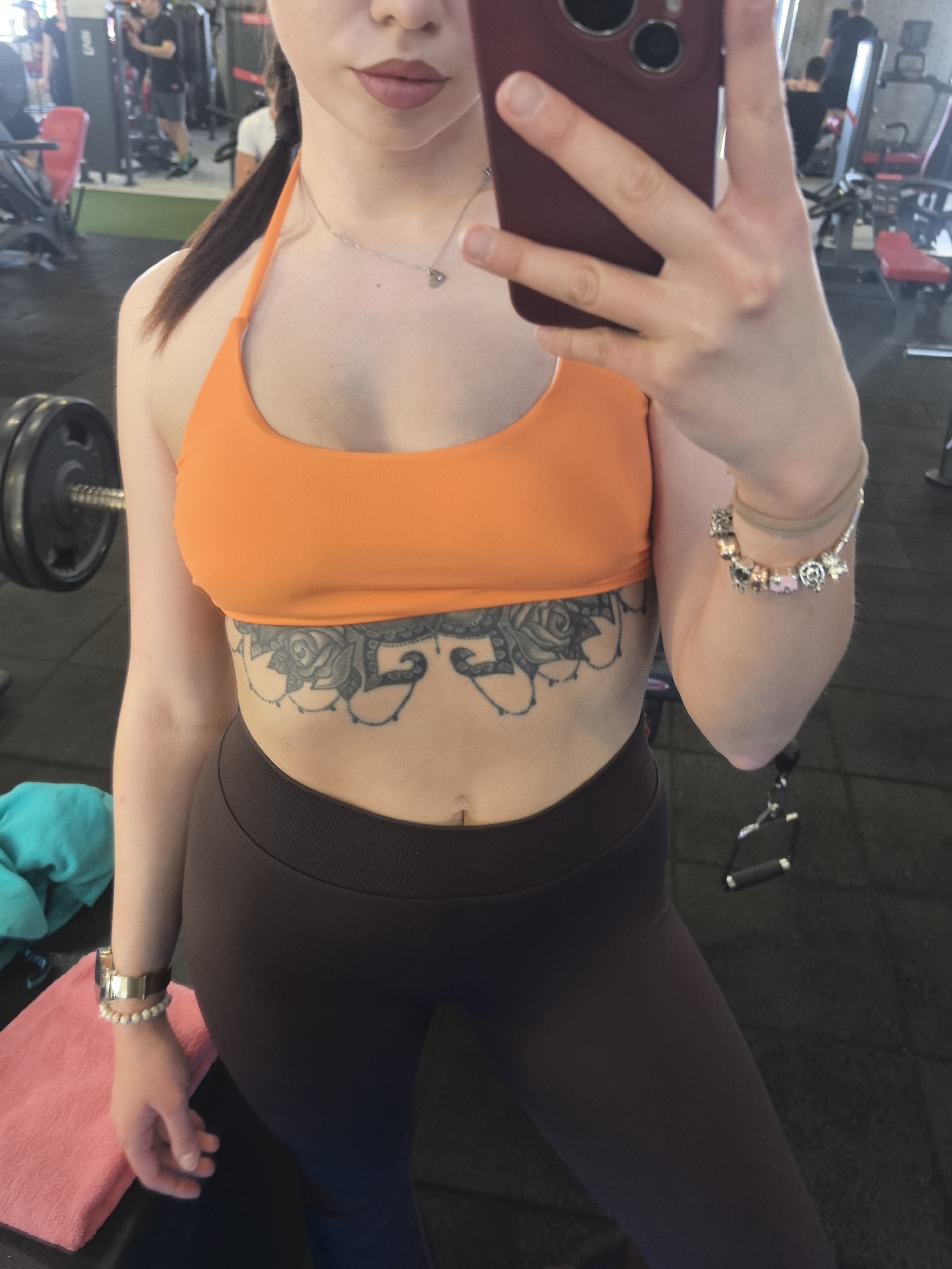 A woman is taking a selfie in a gym. She is wearing an orange sports bra, dark leggings, and has tattoos on her lower abdomen. She is also wearing bracelets on both wrists. In the background, other people are working out on gym equipment. Based on provided information, the location is a gym in Stara Zagora, Bulgaria.