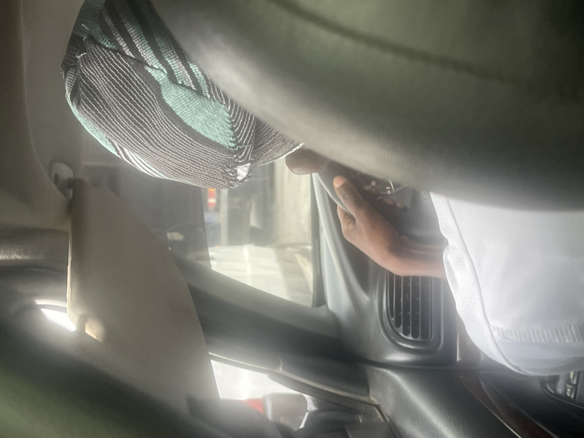The image captures the interior of a vehicle during the daytime, rotated 90 degrees clockwise from its original orientation. A person's dark-skinned arm and hand are visible, gripping a dark-colored steering wheel, indicating they are driving. The driver is wearing a white garment, possibly a traditional robe, a portion of which is seen on the right side of the frame.
On the upper left, a patterned fabric is visible, likely part of the driver's attire or a head covering. It features distinct horizontal stripes in shades of green, black, and white. Parts of the car's dashboard, including air conditioning vents, are discernible below the steering wheel. Bright light streams in through the windshield, suggesting clear, sunny weather outside. The overall scene is bright due to the external light contrasting with the car's dark interior components. No text is visible in the image. This scene takes place in Makera, Nigeria.