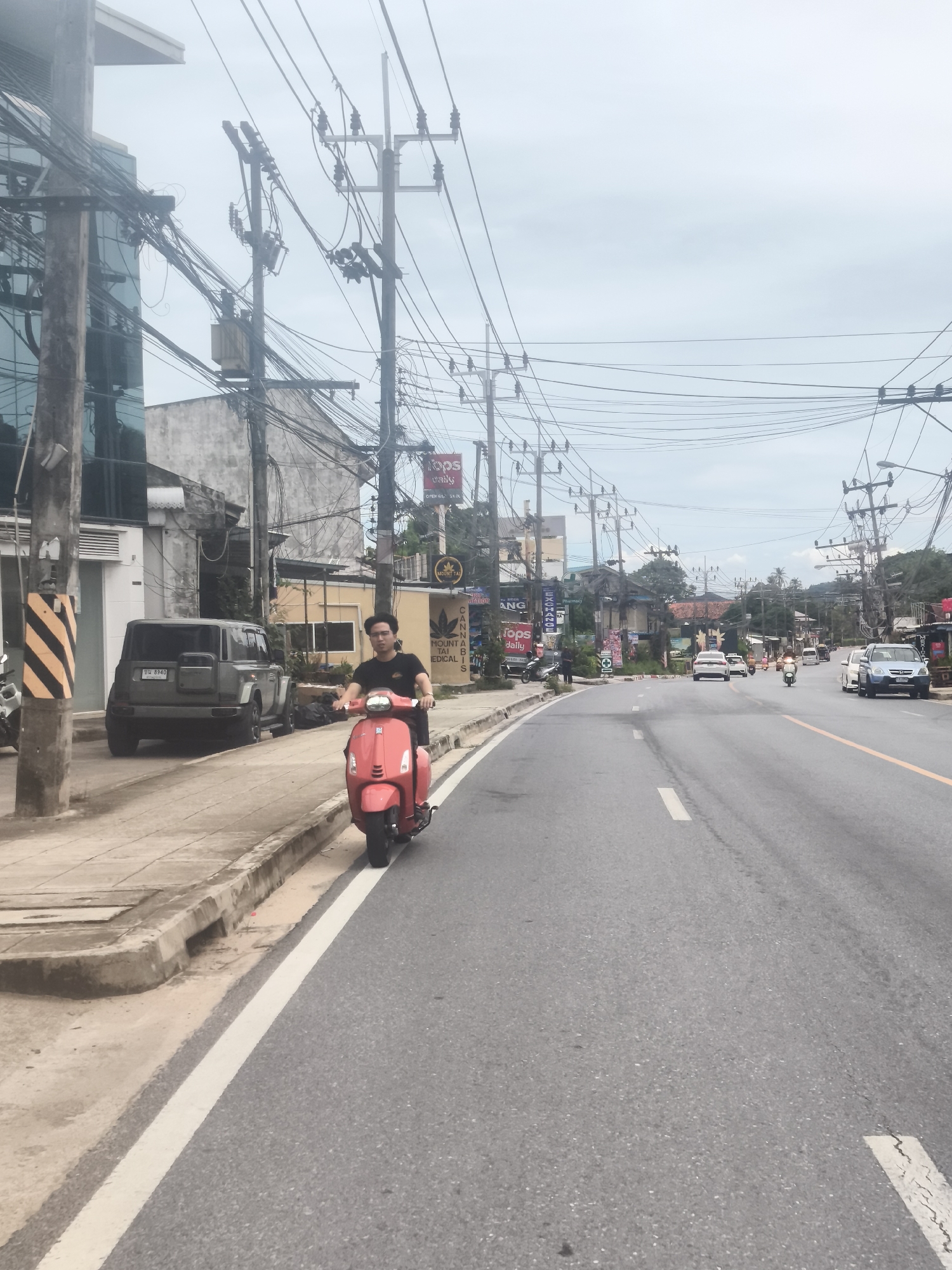 A man is riding a pink scooter on a road in Baan Chaweng Noi, Thailand. The man is wearing a black t-shirt and black shorts. He is looking forward. The scooter is moving forward. The road is paved and there are cars and other scooters on the road. There are buildings on the side of the road. There are also many power lines and utility poles. The sky is overcast. The overall scene is that of a busy street in a tropical location.
