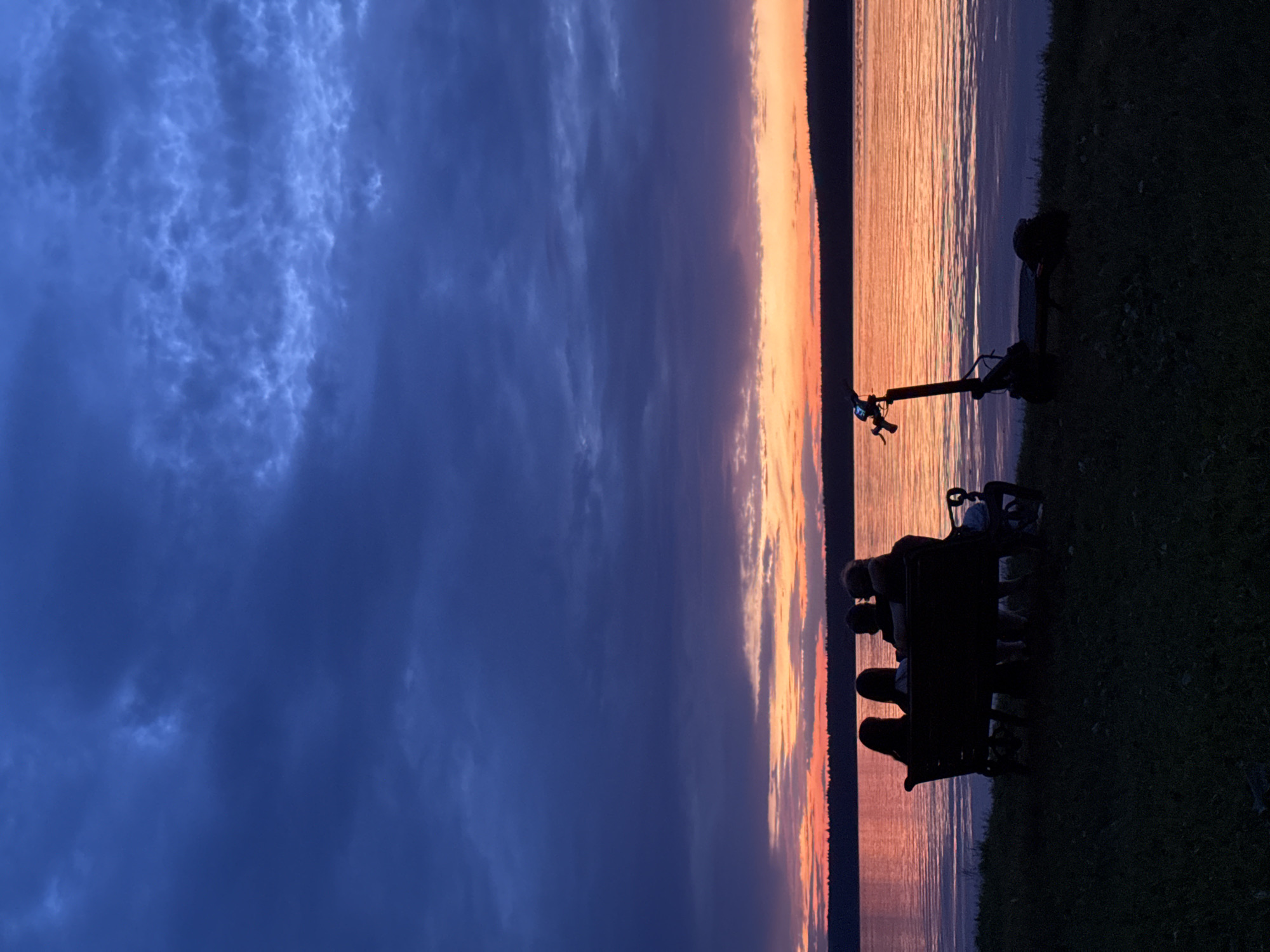 A serene dusk scene unfolds along the shore of a tranquil body of water, possibly a lake or a wide river, with the distant horizon marked by a dark, silhouetted tree line. The sky is a dramatic canvas of deep blues and purples, transitioning into vibrant oranges and pinks near the horizon where the sun has set. These colors are reflected on the rippling surface of the water, creating a shimmering effect.
In the foreground, a dark park bench is occupied by a group of four individuals, silhouetted against the colorful sky, facing the water. Their postures suggest a shared moment of quiet contemplation or conversation. Beside the bench, a black electric scooter stands upright, its handlebars visible. The ground beneath them is a grassy slope, also rendered in shadow. The overall atmosphere is one of peacefulness and natural beauty, capturing a moment of shared experience at twilight. The specific location of Votkinsk, Russia, is not visually ascertainable from the image itself.