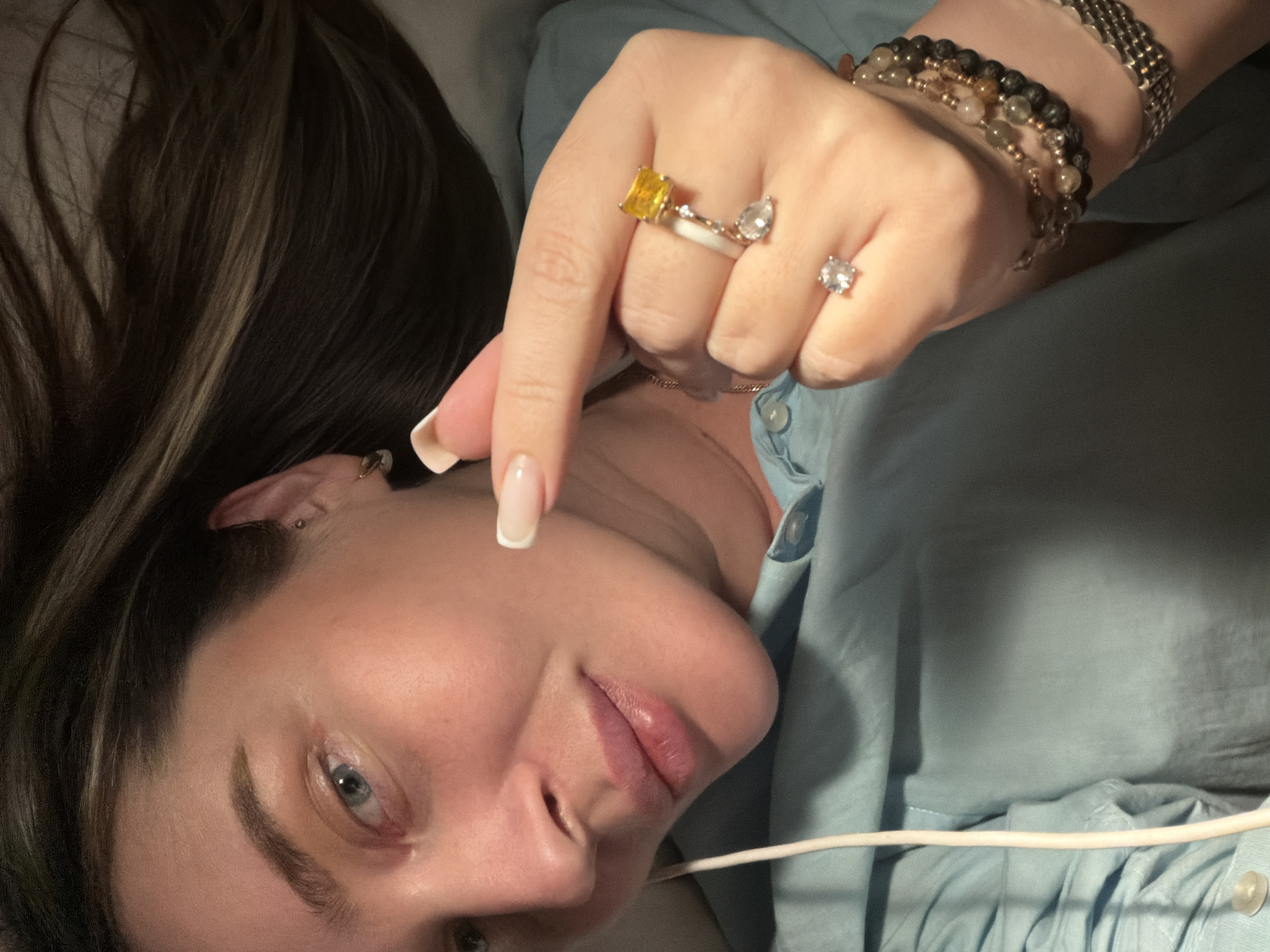 A woman with long, dark hair and fair skin is lying down, facing the camera. She has light blue eyes and full lips. She is wearing a light blue button-down shirt. Her left hand is raised, displaying multiple rings, including one with a yellow stone, one with a teardrop-shaped stone, and a solitaire diamond ring. She is also wearing several bracelets. A white tube or cord is visible near her face. There is no indication of the location being Moscow, Russia.