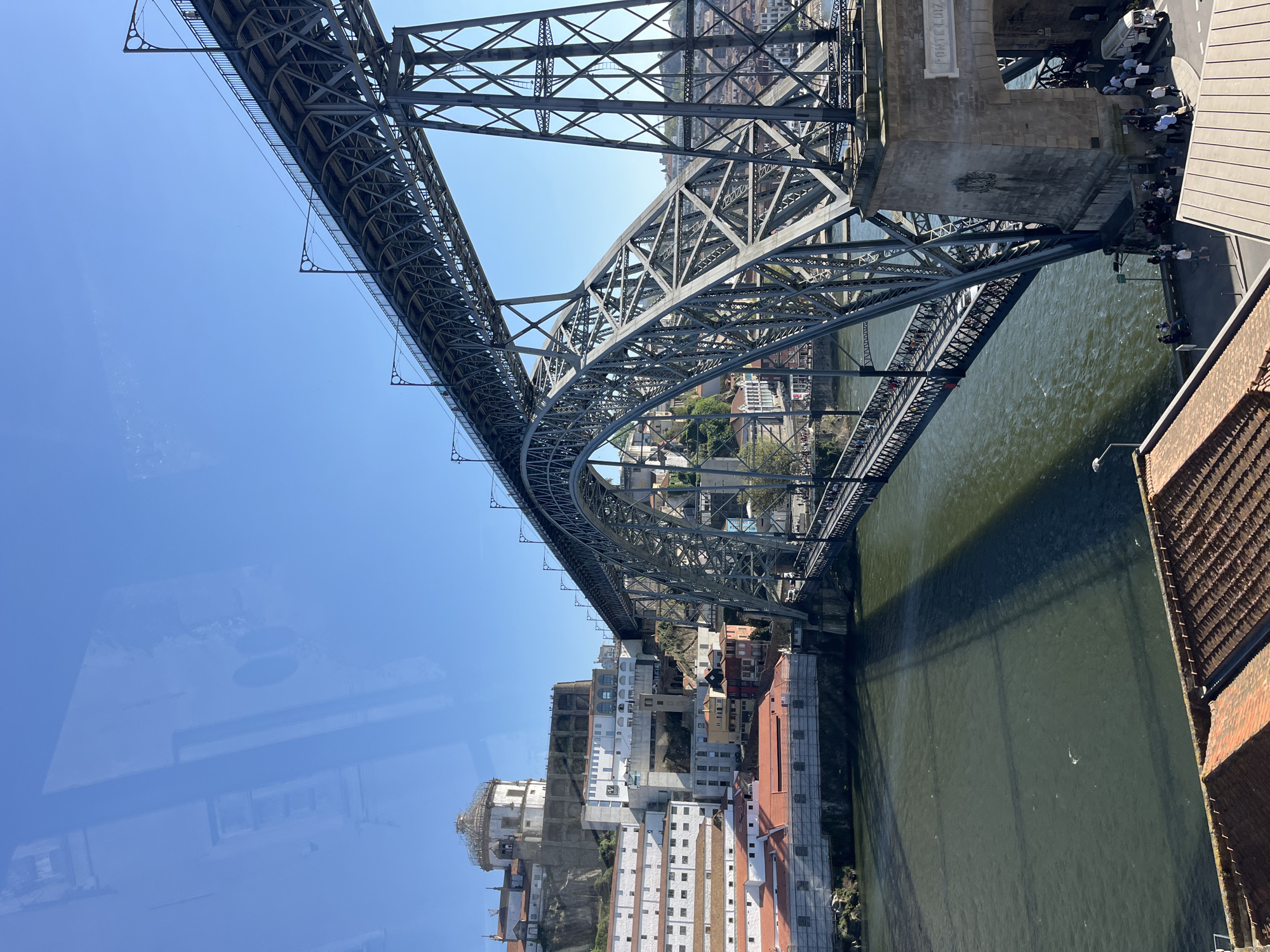 The image shows the Dom Luís I Bridge in Porto, Portugal, from an elevated perspective. The bridge's metal structure arches across the Douro River, with the city's buildings visible along the riverbanks. People are visible on a pedestrian walkway of the bridge. The river appears murky, and a shadow of the bridge falls on the water's surface. The sky is clear and blue. There is a reflection of the surrounding building visible on the left side of the image.