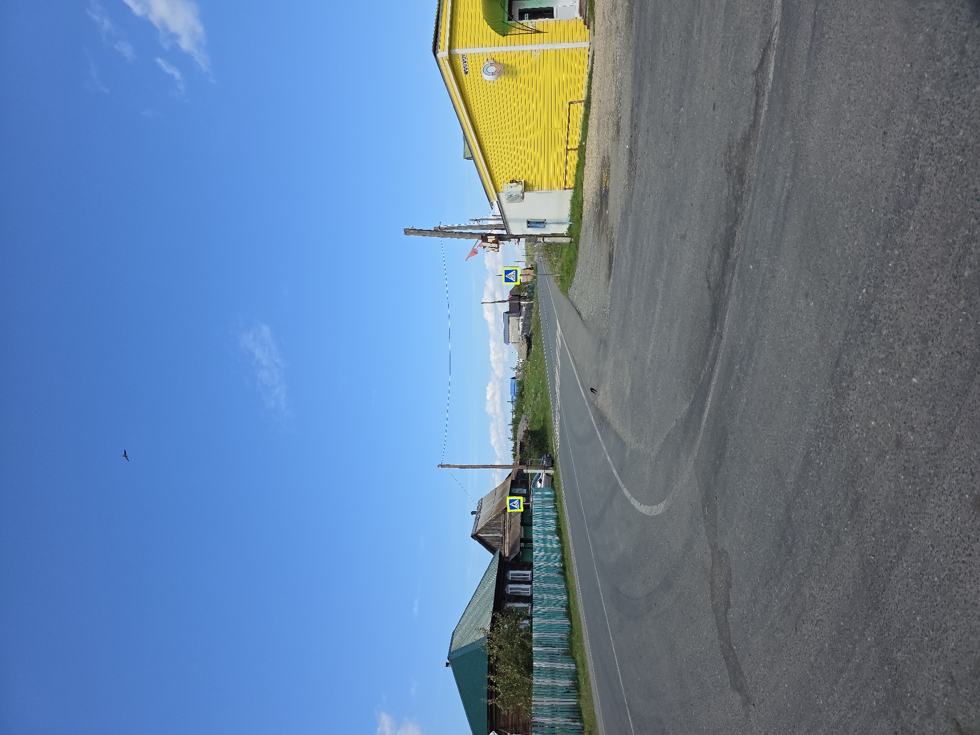 The image shows a rural scene in Khalitova, Russia, on a clear, sunny day. The foreground features a paved road that curves to the left. To the left of the road, there are two traditional wooden houses with green roofs and a white and teal picket fence. A pedestrian crossing sign is visible near the houses. To the right of the road, there is a bright yellow building with a white section and an air conditioning unit. A smaller white building is attached to the yellow building. A wooden utility pole with wires and a small red and white flag is located between the road and the buildings. In the distance, more rural buildings and some clouds are visible under a bright blue sky. A bird can be seen flying high in the sky, and another bird is on the road.