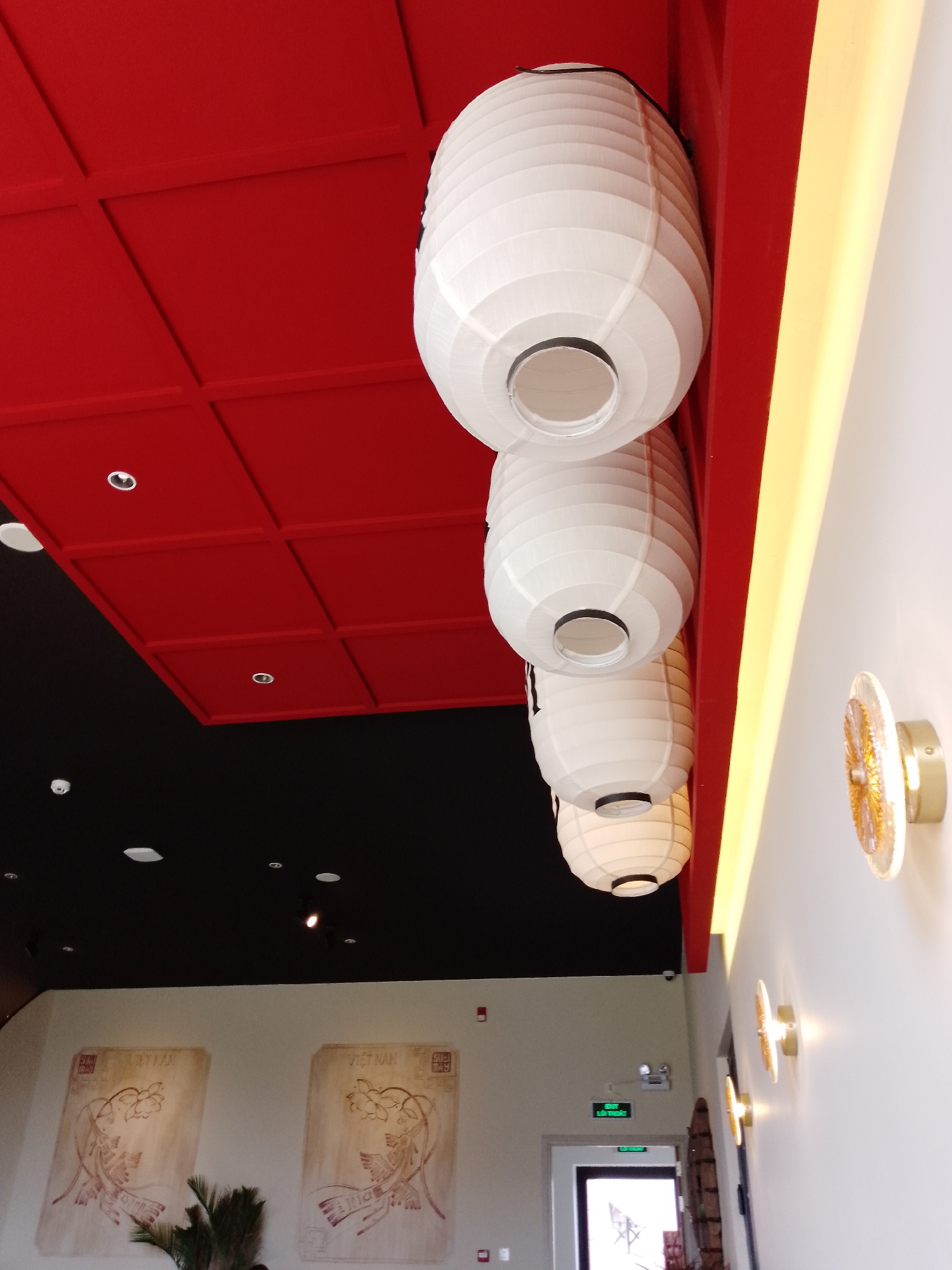 The image displays the interior of a modern space, likely a restaurant or commercial establishment, with a distinct Asian-inspired aesthetic. The ceiling is a prominent feature, divided into sections: a vibrant red portion with a grid of square recessed panels and a contrasting black section also fitted with recessed lights.
Four white, pleated paper lanterns of varying sizes hang vertically from a red beam on the ceiling, creating a focal point in the upper right. Adjacent to these lanterns, a continuous yellow light strip illuminates the intersection where a white wall meets the red ceiling. Along this white wall, several circular, decorative wall-mounted lights with golden-brown, textured surfaces add ambient lighting.
In the background, two large, rectangular wall panels are visible, each adorned with elegant, light brown illustrations of floral motifs. The word "VIETNAM" is clearly printed at the top of each panel, providing a strong geographical and cultural cue. A green plant or foliage partially obscures the bottom left of these artworks. To the right, an open doorway reveals a brightly lit area beyond. Above this doorway, a green sign clearly reads "EXIT LỐI THOÁT," with "LỐI THOÁT" being Vietnamese for "Exit." A security camera is mounted above the door, and a red fire alarm pull station is visible on the wall nearby.
The overall scene is well-lit by a combination of natural and artificial sources, suggesting an indoor setting without indicating a specific time of day. The combination of Vietnamese text, lanterns, and color scheme indicates a themed interior.