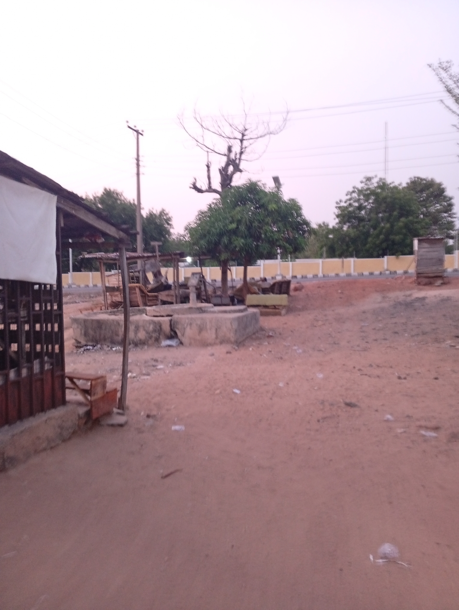 The image depicts an outdoor scene in Zango-ndaji, Nigeria. The foreground is dominated by a dusty, reddish-brown ground with scattered debris. A building with a weathered wooden structure and partial metal siding is on the left. Further back are what appears to be partially burnt wooden structures on concrete foundations. Behind those are trees, a cream and yellow wall, and a wooden outhouse-like structure. An utility pole is visible. The sky is overcast. There are no people visible.