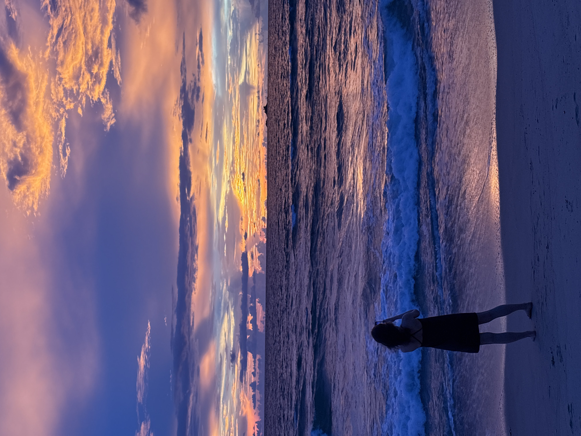 A silhouette of a woman with dark, curly hair, wearing a black dress, stands on a beach facing the ocean. She is holding a phone up, seemingly taking a picture or video. The ocean has waves crashing onto the shore, with the water appearing a deep blue. The sand next to her is a pale, reflective surface.
The scene is set during a sunset. The sky is filled with dramatic clouds, tinged with vibrant orange, yellow, and pink hues, contrasting with the deep blue of the sky and ocean. The sun appears to be low on the horizon, casting a warm glow across the sky and reflecting on the water and sand.
The overall mood of the image is peaceful and serene, capturing a moment of solitary appreciation for the natural beauty of the sunset over the ocean. The location is identified as Choeng Thale, Thailand. There is no visible text in the image.