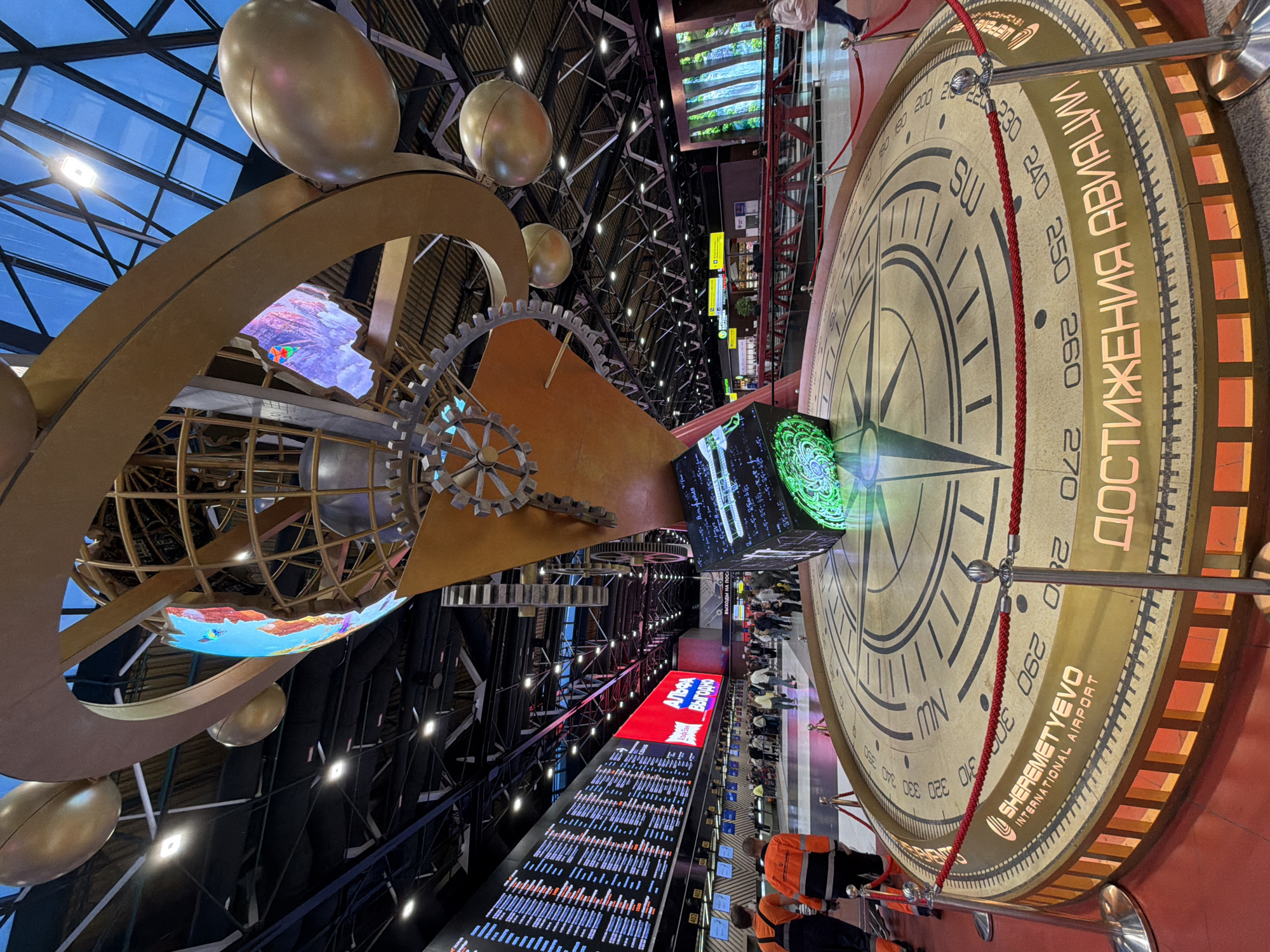 This photograph shows the interior of Sheremetyevo International Airport, which is located in the city of Khimki, Russia. The view is angled upwards toward the airport ceiling and focuses on several artistic installations.
One large artistic installation features a compass rose on the right side of the image with labels in both English and Russian. It reads "SHEREMETYEVO INTERNATIONAL AIRPORT" and displays directions like NW and SW along with numerical degree markings. A red rope barrier surrounds the compass.
On the left, another installation features a series of golden spheres, rings, and gears, along with a digital display inside a sphere-shaped structure. The background shows the airport's ceiling structure with lights, and in the middle distance, there are flight information display boards with various data.
A red sign displaying the word "АЛЬФА" and "Выгодно" in Russian is shown above the flight information boards. People in airport worker vests are visible in the foreground, and there are other passengers and airport employees in the background, implying that people are waiting for flights or working.