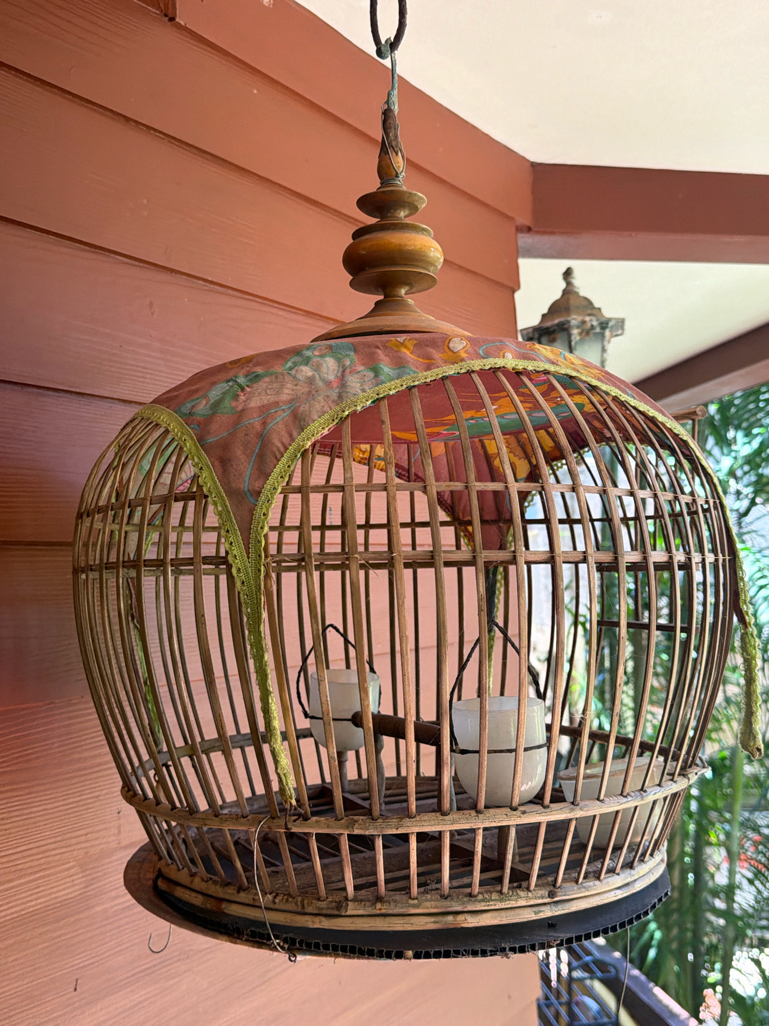This is a close-up, eye-level, medium shot of a decorative bamboo birdcage hanging from an overhang. The birdcage is circular with a domed top, adorned with a fabric covering featuring a floral pattern. Inside the cage, two white cylindrical objects, possibly candle holders or feeders, are suspended. The background features a wooden-paneled wall on the left and lush green foliage on the right, suggesting an outdoor or semi-outdoor location in a tropical setting like Ko Samui District, Thailand. A light fixture is visible in the upper right corner. The overall impression is one of traditional craftsmanship and an outdoor ambiance.