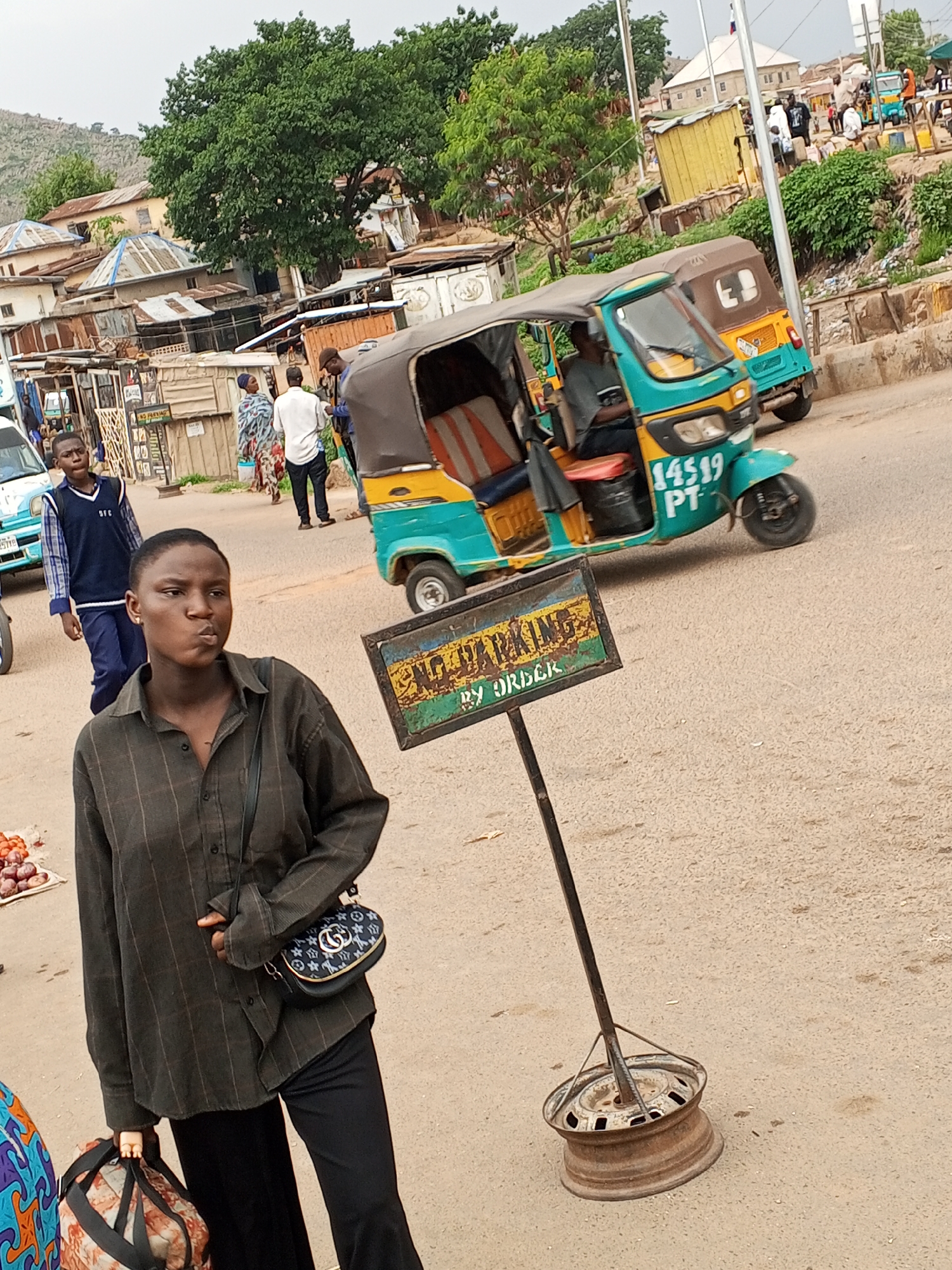 The image depicts a street scene in Gwong, Nigeria. In the foreground, a young woman is walking towards the viewer. She is wearing a dark, button-down shirt, black pants, and a small purse over her shoulder. She is carrying a large patterned bag in her left hand and appears to be chewing something.
Behind her and to the right stands a "No Parking By Order" sign, affixed to a metal pole and stabilized by a rusted car wheel rim at the base. Behind the sign, a blue and yellow motor rickshaw is in motion, with a driver visible inside.
In the background, various buildings with corrugated metal roofs can be seen, along with a mix of pedestrians and vehicles. Other parked motor rickshaws are also present. The environment suggests a bustling urban area, possibly a market or commercial district.