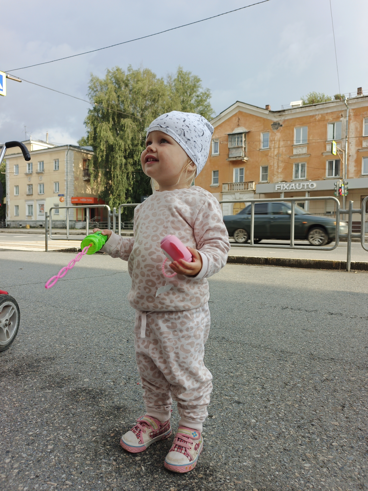 A full-body, eye-level outdoor shot captures a young Caucasian child standing on an asphalt road. The child, wearing a light-colored beanie with a patterned design and a matching light-pink sweat suit with a leopard-like print, is holding two toys: a green bubble wand in their left hand and a pink object, possibly a toy phone, in their right. The child's gaze is directed upwards and to the left, with a slight smile. Behind the child, a silver stroller is partially visible on the left side of the frame. The background reveals a city street scene in Zlatoust, Russia. Two multi-story buildings with orange and yellow facades line the street, featuring numerous windows and balconies. One building displays a sign reading "FIXAUTO." A dark green car is in motion on the road, blurred due to movement. Metal barriers and a traffic light with a red signal are also present. The sky is overcast with gray clouds.