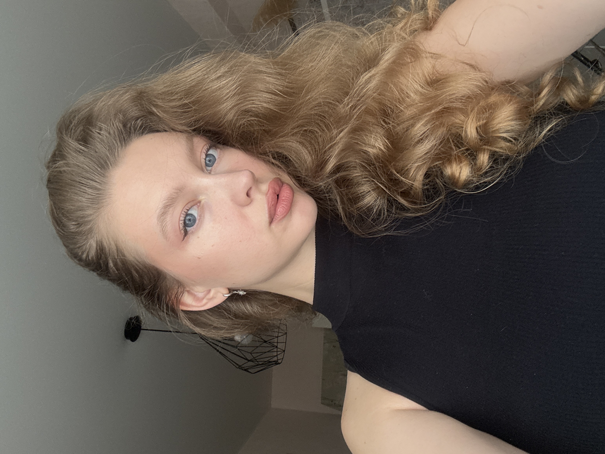 A young woman with blonde, curly hair is pictured indoors, possibly in her home in Izhevsk, Russia. She is wearing a black sleeveless top and a silver earring. Her makeup includes subtle eyeliner and lipstick. In the background, a black geometric pendant light fixture is visible hanging from the ceiling. The lighting suggests it may be daytime.