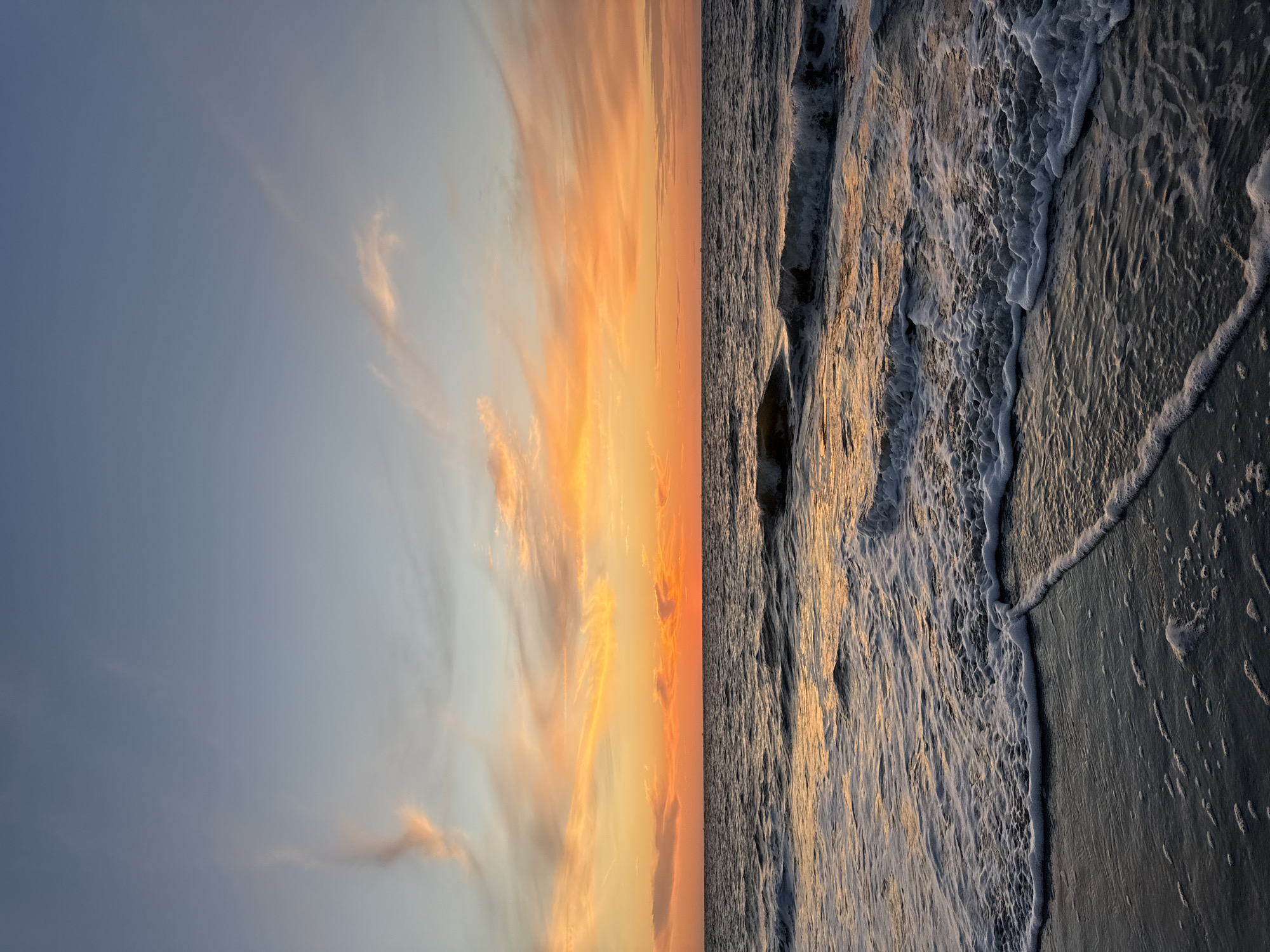The image captures a sunset over the North Sea on the island of Langeoeg, Germany. The sky is a gradient of blue at the top, transitioning to vibrant orange and yellow hues near the horizon, illuminated by the setting sun. Wispy clouds are scattered across the sky, catching the warm light. The sea below is dark and choppy, with waves creating white foam as they approach the shore. The wet sand in the foreground reflects the colors of the sunset. There are no people or identifiable objects visible in the scene, only the natural elements of the ocean, sky, and beach.