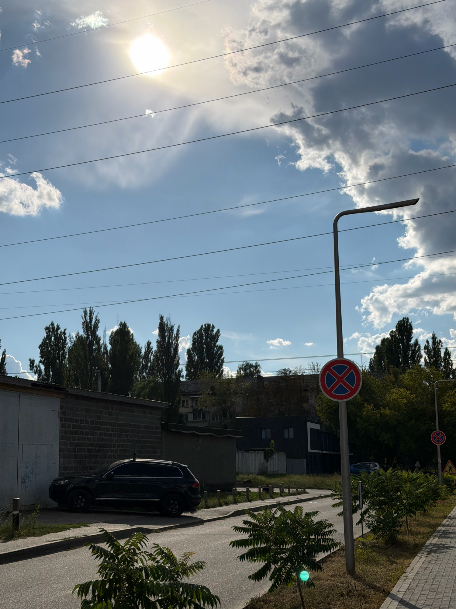 The image depicts a street scene in Kyiv, Ukraine. The sun shines brightly through a cloudy sky with several power lines crossing the frame horizontally. On the left, a dark-colored SUV is parked in front of a brick building with a large, white garage door. A street light with a "no stopping" sign is visible on the right side of the road, along with a pedestrian sidewalk made of paving stones. Lush green trees and bushes line the sidewalk. In the background, several tall, slender trees stand against the sky, and various buildings are visible. A smaller traffic sign and a green traffic light are also present.