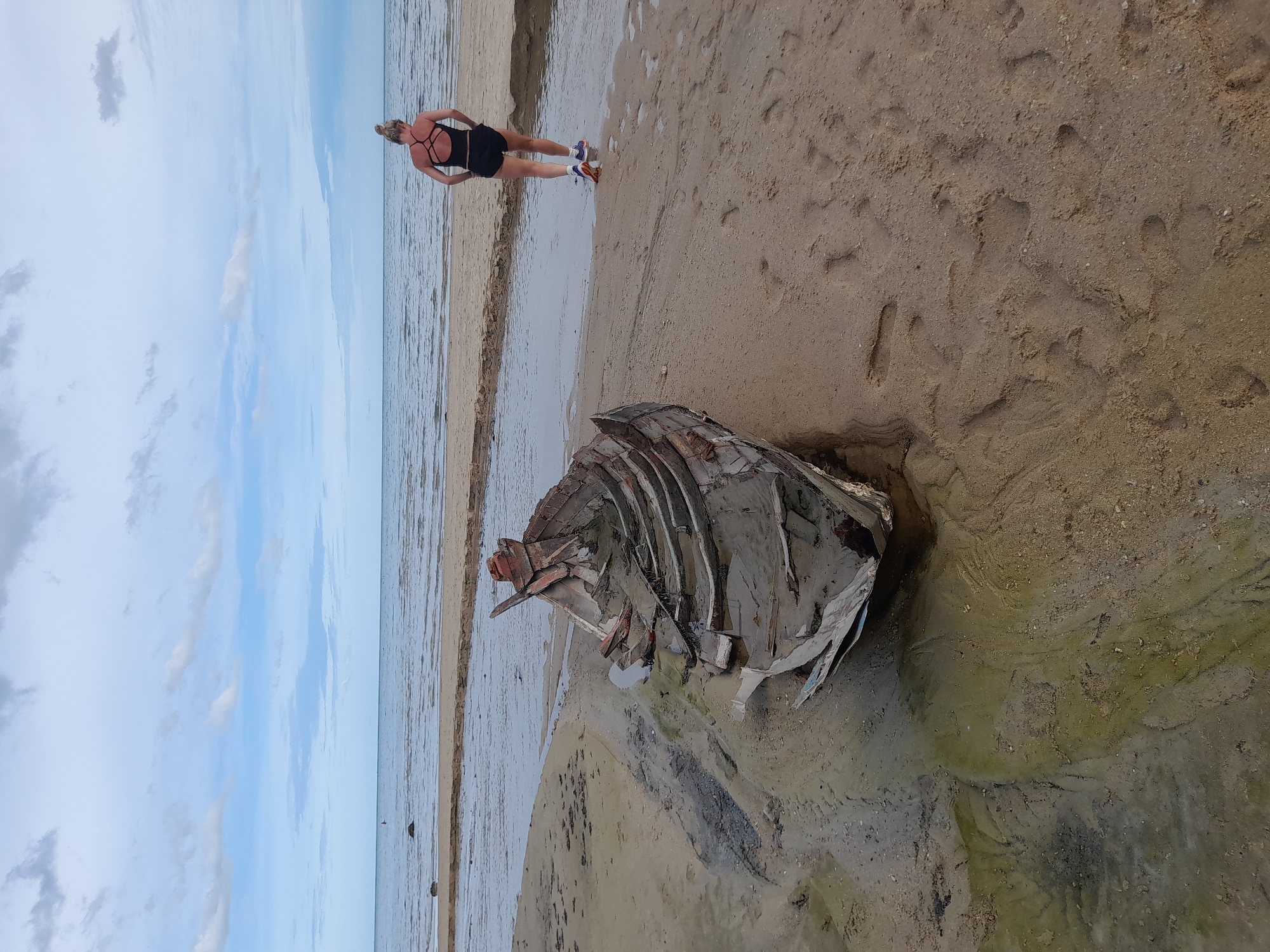 An overhead shot shows a person walking on a sandy beach with a partially submerged shipwreck in the foreground. The person, facing away from the camera, is wearing athletic clothing, including black shorts and a black tank top with a criss-cross back. They are also wearing white socks and athletic shoes with blue and orange accents. The shipwreck is old and appears to be made of wood, with its ribs exposed. It is partially filled with water and sand, and some debris is scattered around it. The beach is wet in some areas, indicating the tide may have recently receded. In the background, the ocean stretches to the horizon, with a calm, rippled surface under a cloudy sky. The sand is light brown and has several footprints visible. The scene appears to be in a natural setting, possibly a coastline, with no urban elements visible.