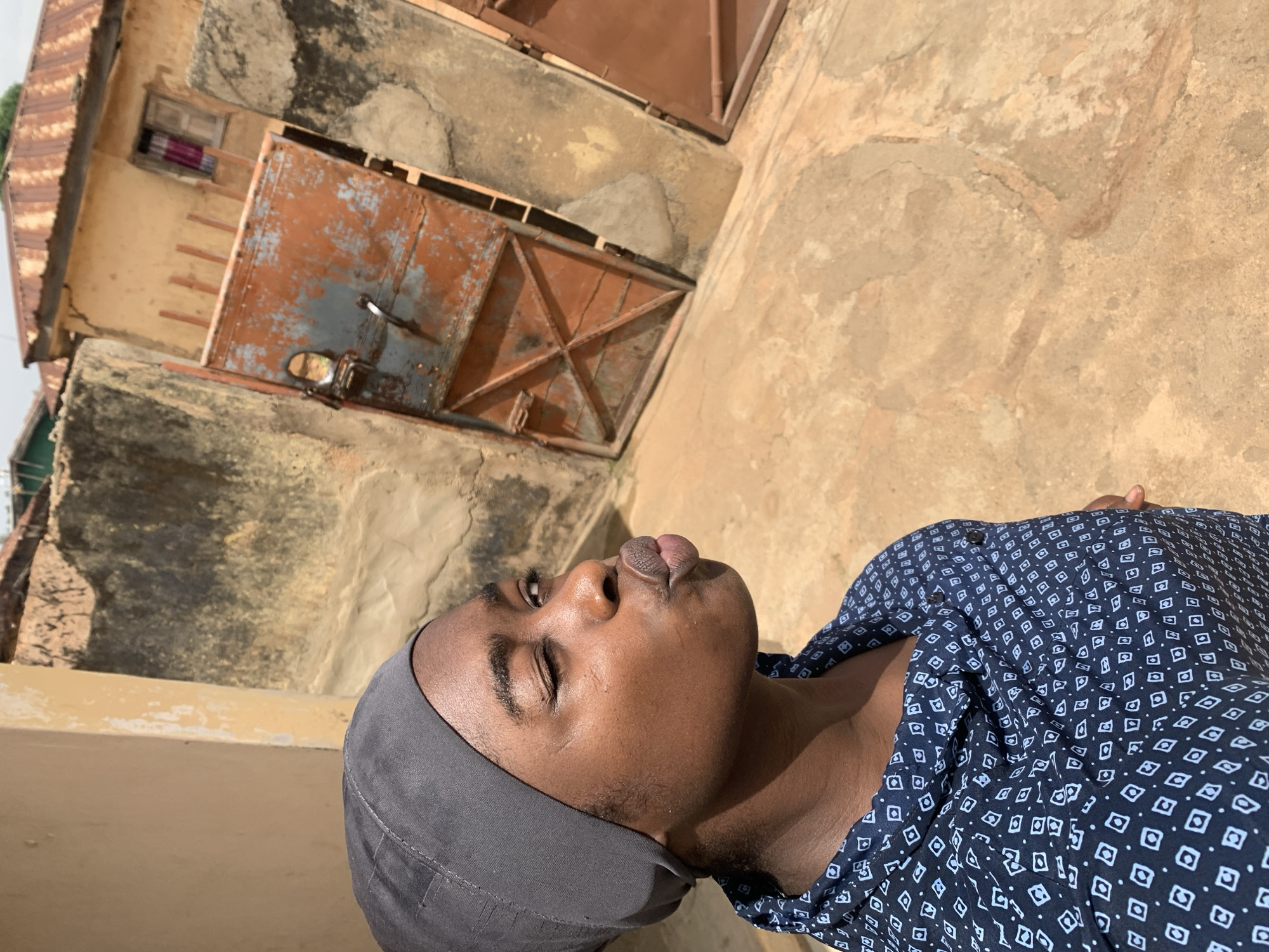 A person wearing a grey head covering and a patterned blue shirt is lying outside near weathered buildings in Rafin Par, Nigeria. The buildings have reddish-brown doors and windows. The person's eyes are closed, and their face is angled toward the sun.