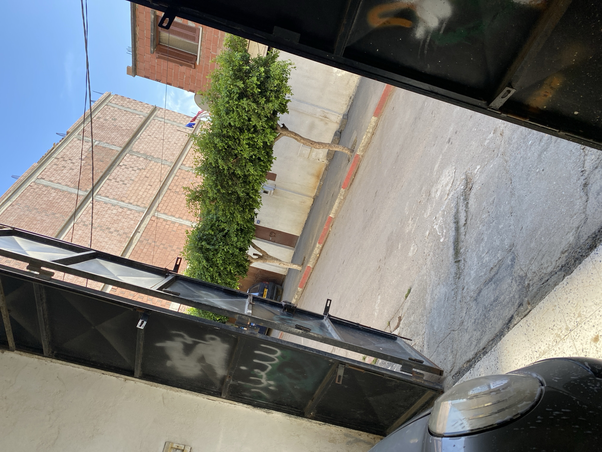 The image is taken from a slightly elevated perspective, looking through an open metal gate towards a street in Baraki, Algeria. The gate is black and has some white and green spray paint graffiti on its left panel. To the left of the gate is a textured white wall with a light switch visible.
The street is paved with asphalt and has a curb lined with red and white paint. On the right side of the street, there is a row of buildings. The closest building appears to be a residential structure with multiple floors under construction, made of exposed brick. Further down the street, another building with red brick and closed shutters is visible.
In front of the buildings are two trees with dense green foliage. A black car with a partial license plate "1065 YN 16" is parked on the street. The sky is clear and blue. The overall impression is of a typical urban street scene in a residential area, possibly with ongoing construction.
