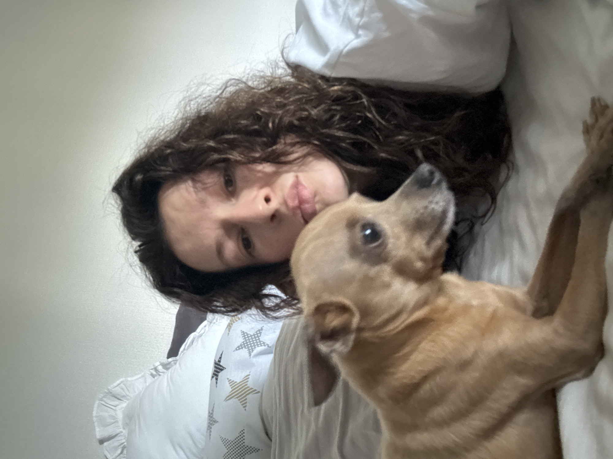 A woman and a tan chihuahua are lying on a bed in what appears to be an indoor setting. The woman has curly dark hair and is looking at the camera with a pouting expression. The dog is in the foreground, facing slightly away from the camera, with one of its paws visible. The bedding includes a pillow with a star pattern and a white comforter. The location in the city of Chaikovsky, Russia is not verifiable from the image alone.