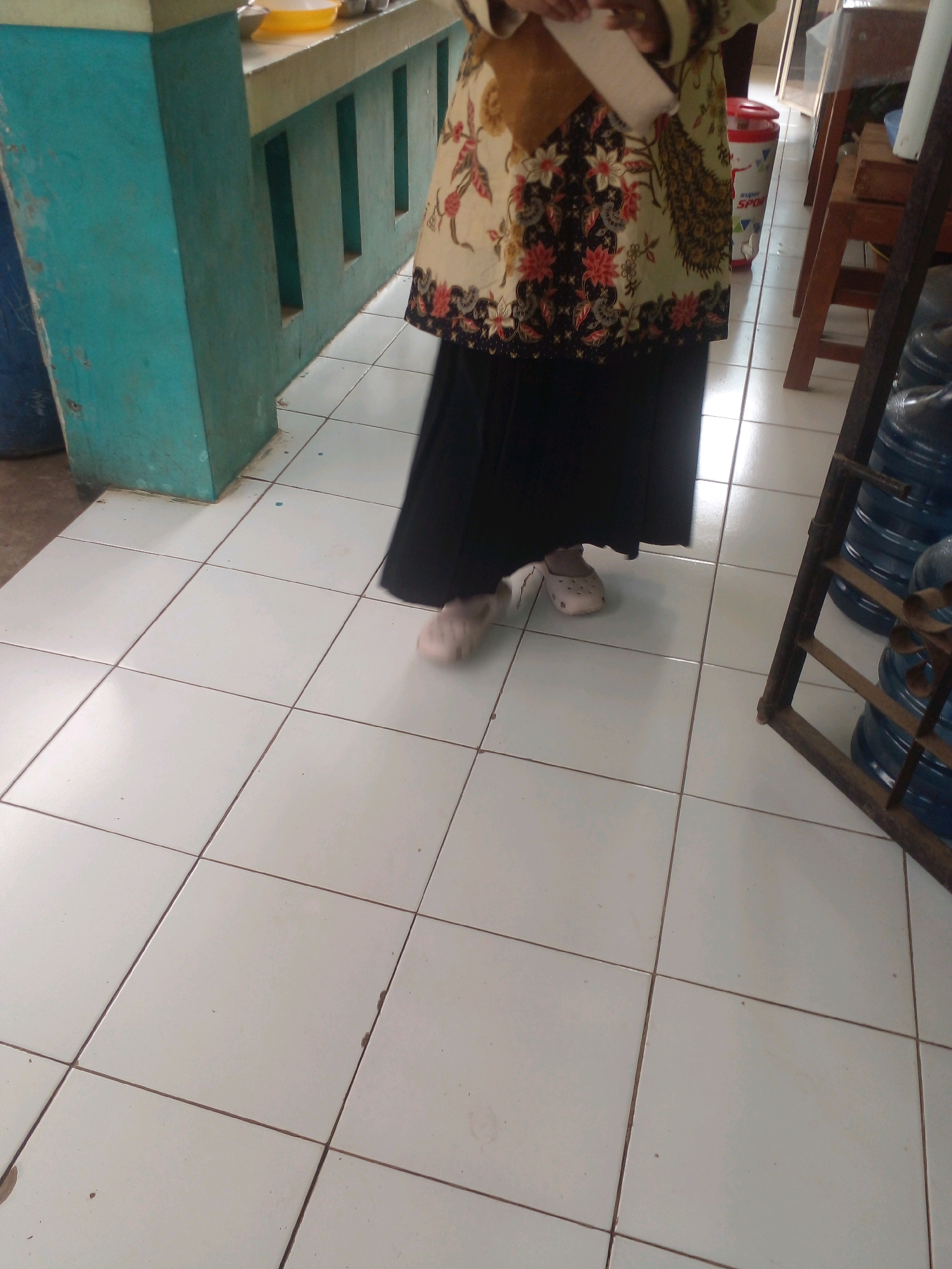 The image depicts a person standing on a tiled floor indoors, likely in Banyuputih, Indonesia. The person is wearing a dark, long skirt and a patterned top with floral and peacock motifs. They are also wearing light-colored clogs. To the left of the person, a teal-colored pillar is visible, which appears to be part of a counter or structure. In the background, there is a red and white cylindrical object, possibly a trash can or dispenser, and several large blue water bottles stacked on a metal rack to the right. The floor is made of square white tiles with dark grout lines. The lighting suggests an indoor environment with ambient light.