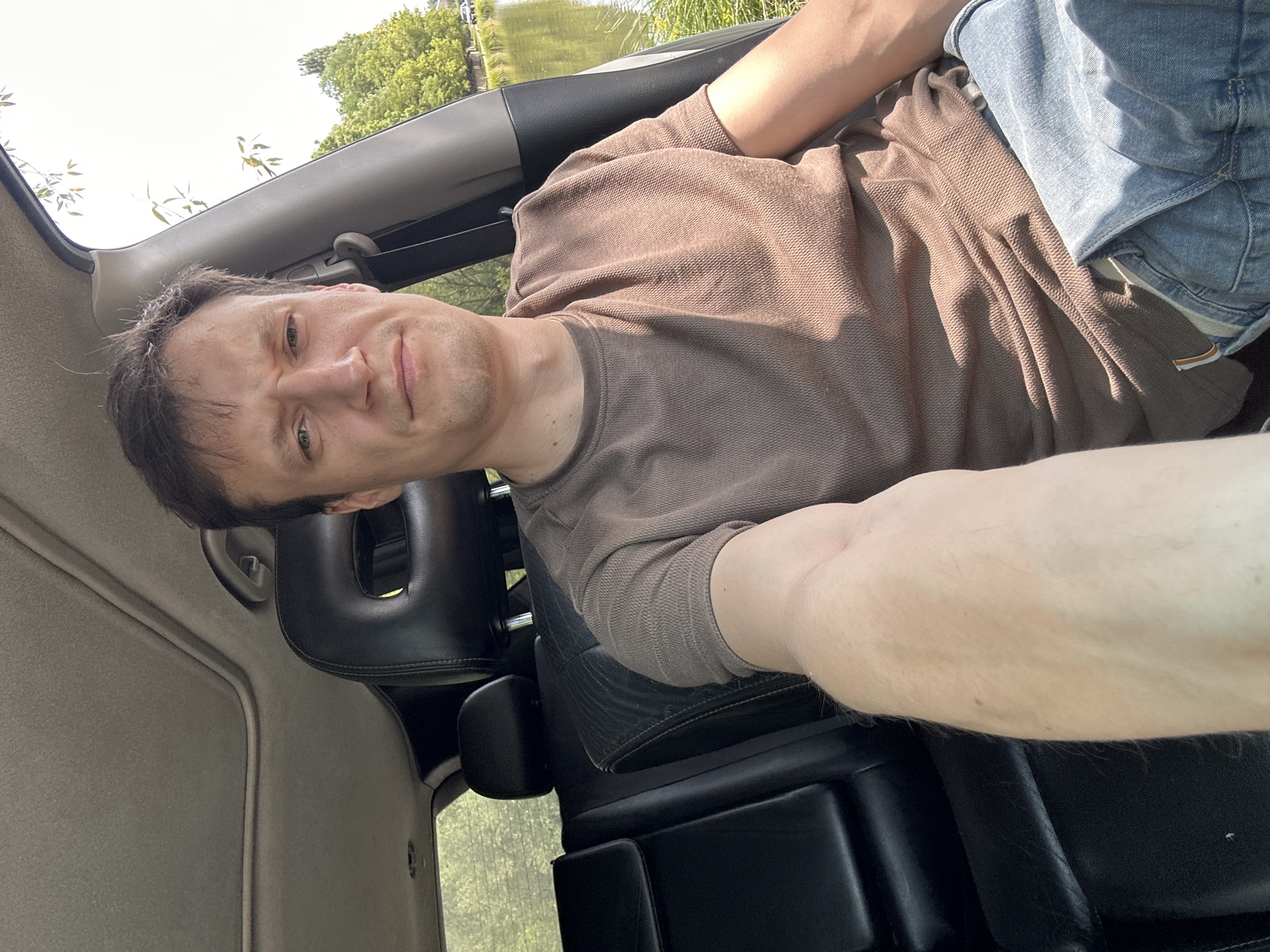 A man is taking a selfie from the front passenger seat of a car. He has brown hair and is wearing a brown t-shirt and blue jeans. The car interior is visible, including black leather seats and a sunroof. Outside the car window, a body of water, likely a river or lake, is visible, with trees and greenery lining the bank. The scene suggests a daytime setting in Makiv, Ukraine, possibly near a natural area.