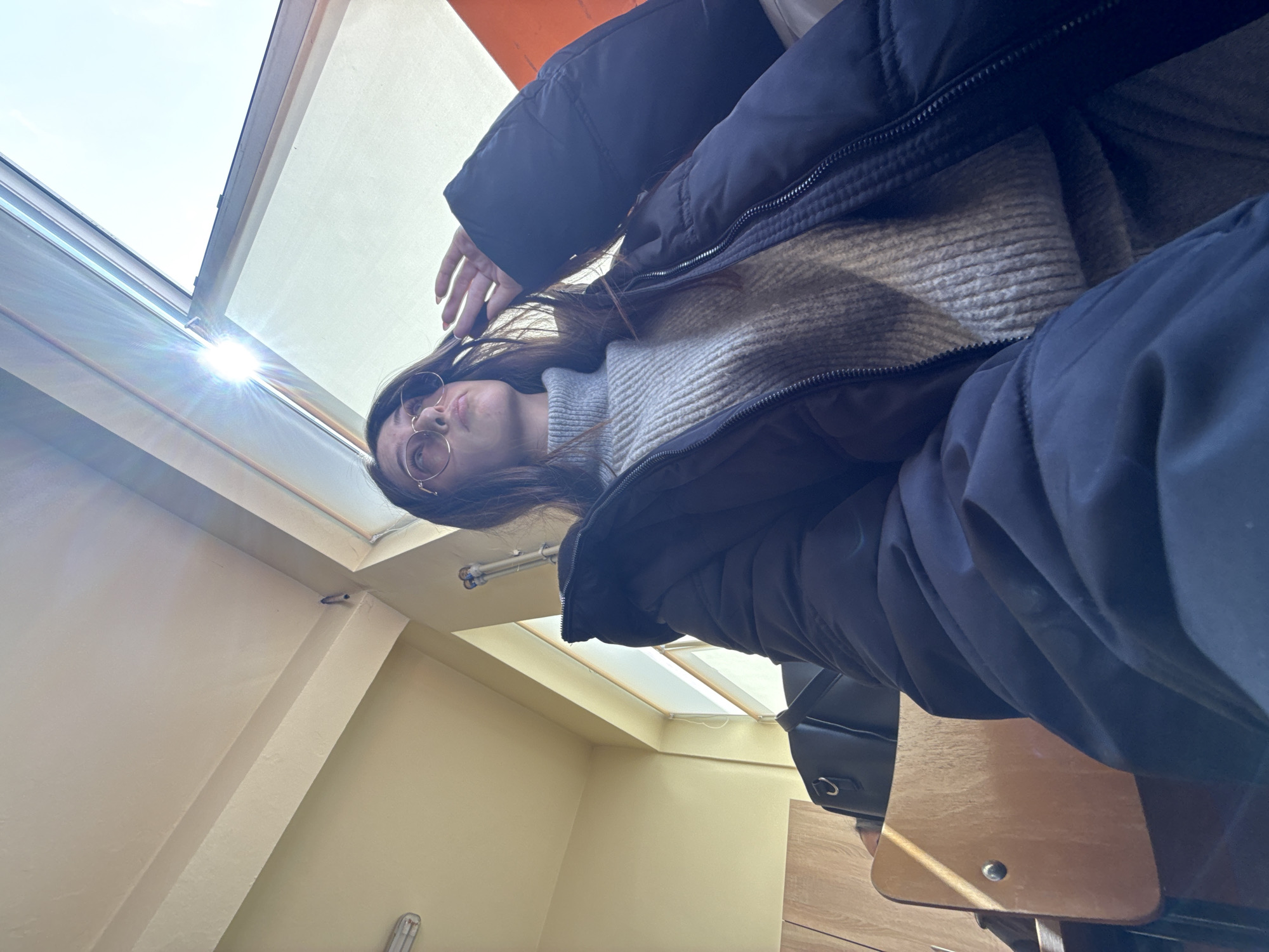 A young woman is the primary subject of this low-angle, indoor photograph. She is wearing round glasses, a grey sweater, and a dark, puffy jacket. Her hand is near her face, seemingly shielding her eyes from the light.
The scene is brightly illuminated by sunlight streaming in from what appears to be a skylight or a high window. The room's walls are painted a pale yellow.
The photo was taken inside a building, with the woman appearing to be seated at a wooden desk or table, indicated by a visible edge. There is also a black object, possibly a bag, hanging near the table.
Given the architecture and available details, it's impossible to confirm that the location is Sofia, Bulgaria. There is no conclusive evidence of the city or country present in the image.