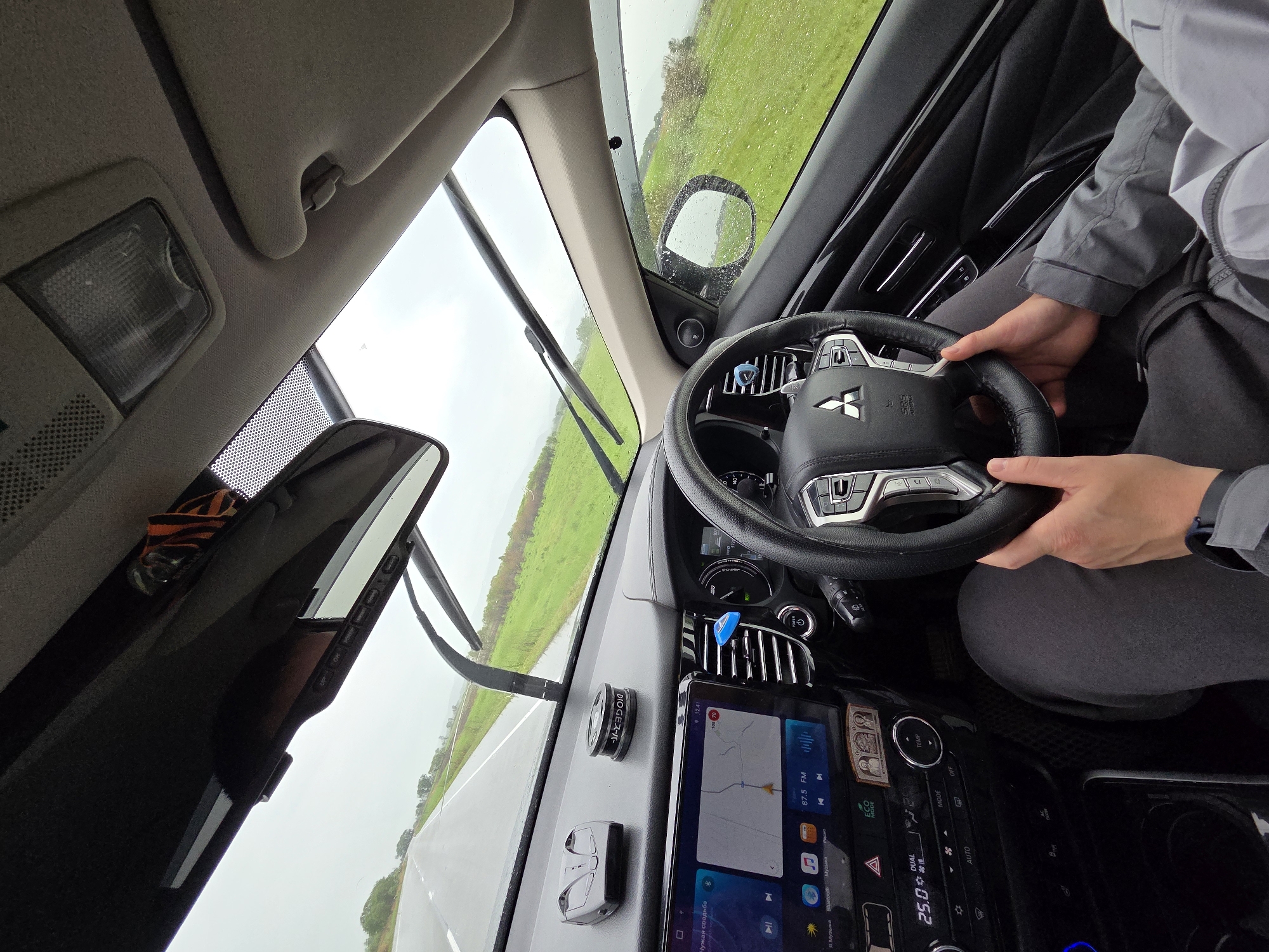 An interior shot of a car shows a driver's perspective on a rainy day in what appears to be Бабстово, Russia. A person, wearing a grey jacket and dark pants, holds the Mitsubishi-branded steering wheel, suggesting they are driving or about to drive. Raindrops are visible on the windshield and side mirror, with the wipers in an upright position, indicating wet weather conditions.
Through the windshield, a green field borders a wet asphalt road, which stretches into the distance under an overcast sky. Inside the vehicle, a large touchscreen infotainment system on the dashboard displays "12:41," a navigation map, and "87.5 FM" radio with the song title "Чужая свадьба" (Someone else's wedding) and "Я. Музыка" (Yandex.Music). The climate control shows "25.0" degrees Celsius, "DUAL," and "ECO MODE." Other visible text includes "MITSUBISHI" and "SRS AIRBAG" on the steering wheel, and "DIOGEスーパー" (DIOGE Super) on a car air freshener/diffuser on the dash. A small blue clip is attached to an air vent, and a dashcam or similar device is visible near the rearview mirror. The overall scene suggests a journey in rural Russia during daytime rain.