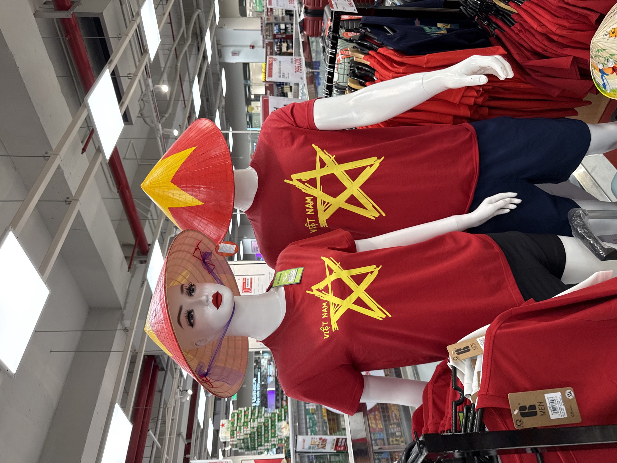 The image shows two mannequins in a retail store, likely a department store or supermarket in Nha Trang, Vietnam. The mannequins are dressed in red t-shirts with a yellow star and "VIỆT NAM" printed on them, and are wearing traditional Vietnamese conical hats (nón lá), the hats are red and yellow. The mannequin on the left is female, with makeup applied to its face, while the mannequin on the right is male. In the background, shelves are stocked with various products, including beverages and clothing. A price tag of 799,000 can be seen on a display in the background. The store appears to be well-lit with fluorescent lighting.