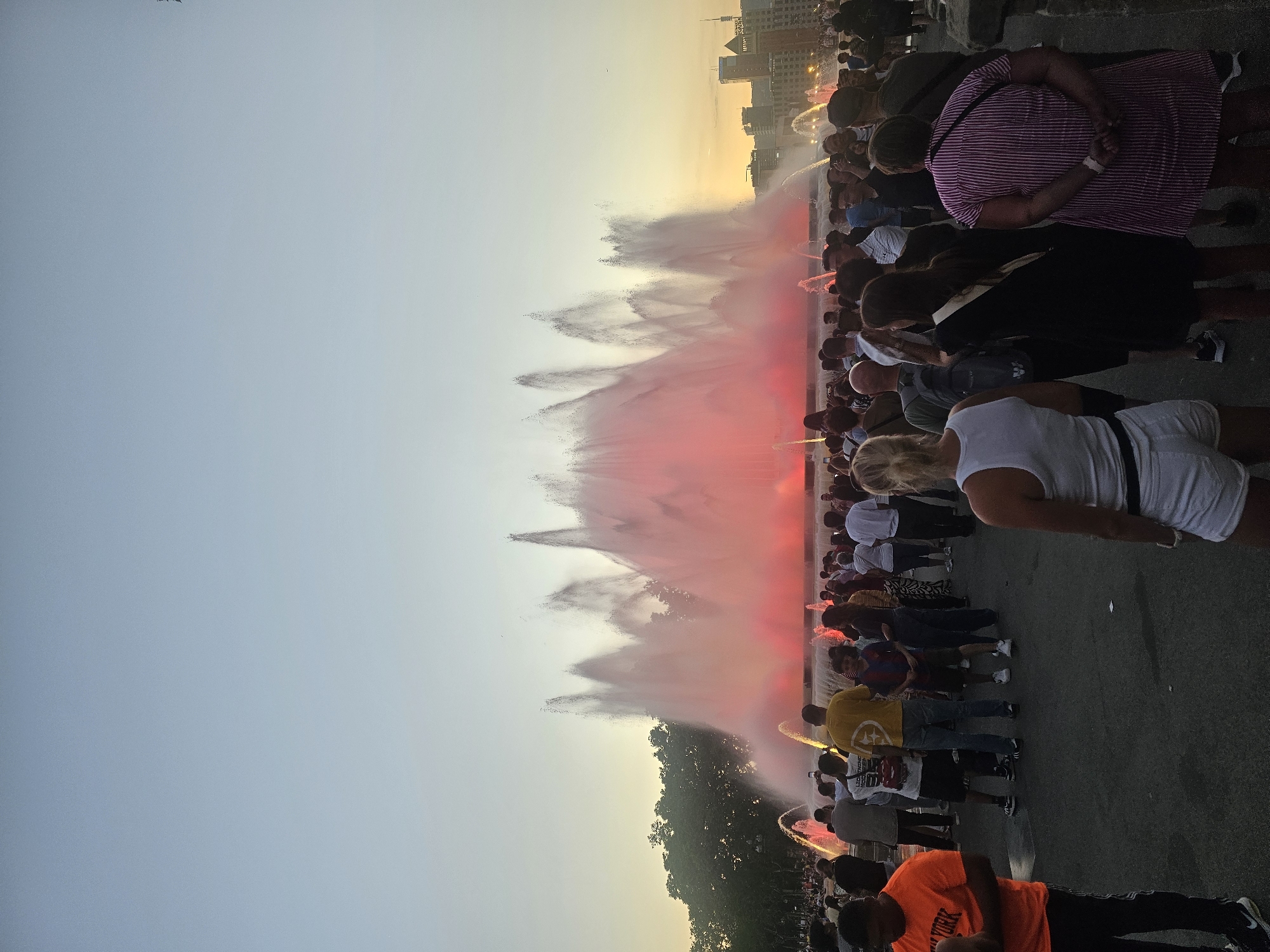 A crowd of people are gathered in front of a large, illuminated fountain in Barcelona, Spain. The Magic Fountain of Montjuïc is in the background, with its water jets shooting high into the air and glowing with red and pink light. The sky above is a soft gradient of light blue and orange, indicating either dawn or dusk. The crowd is diverse, with people of various ages and ethnicities. Some are looking at the fountain, while others are taking photos or videos with their phones. The ground is paved with asphalt. The architecture of buildings can be seen in the distance behind the fountain.
