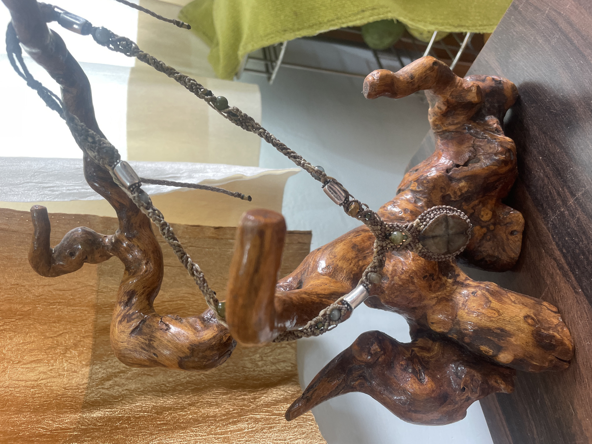 A necklace with a stone pendant is displayed on a wooden sculpture. The sculpture is abstract and organic in shape, with multiple branches and twists. The necklace is made of braided cord and features several small beads and a larger, flat, faceted stone as the pendant. The background consists of textured fabric in shades of gold and off-white, and a glimpse of a green textile on the right. The setting appears to be an indoor display. The image is a medium shot, focused on the necklace and the sculpture.