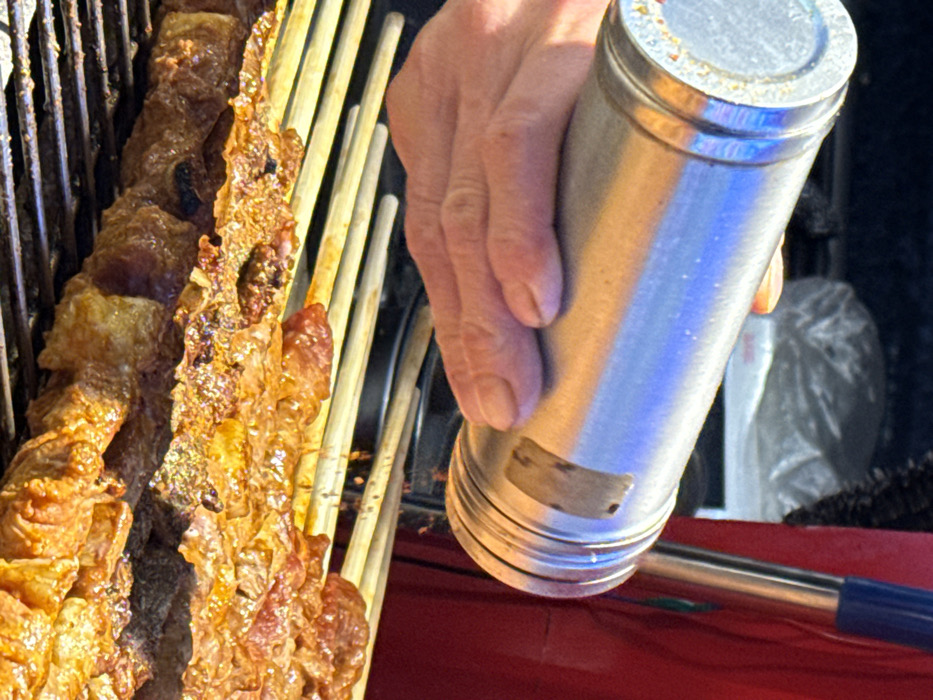 The image depicts a close-up, high-angle view of food preparation taking place in Yuzhong District, China. A human hand, exhibiting fair skin, holds a metallic, cylindrical spice shaker with a perforated top, tilted downwards. The shaker reflects hues of blue and orange from ambient light and has a small, light-brown adhesive patch on its side. It is actively dispensing dark, granular particles, consistent with spices or seasoning, onto several skewers arranged below. These skewers, made of light-colored wood, rest horizontally on a metal grill grate. Each skewer holds multiple pieces of reddish-brown meat, which appear cooked or partially cooked, some displaying charred edges and a visible coating of seasoning. To the right of the frame, a section of red fabric, possibly clothing or an apron, is visible, alongside a partial blue handle of an unknown utensil. The background, largely out of focus, shows a white plastic bag containing an indistinct object against a dark surface. The scene suggests a street food or market stall environment.