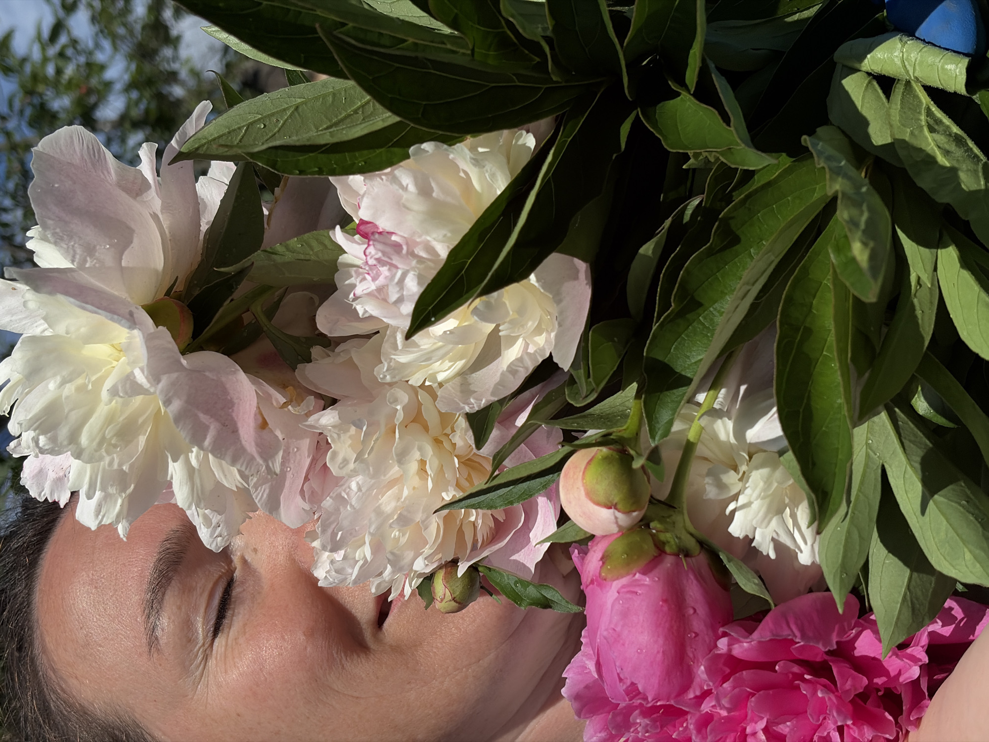 In a close-up shot, a woman's face is partially obscured by a vibrant bouquet of peonies. Her eyes are closed, and she appears to be smelling the flowers, her lips slightly parted. The peonies are a mix of white, cream, and pink hues, with some buds still closed. Lush green leaves surround the flowers. The lighting suggests it is daytime, possibly outdoors, with the sky visible in the upper right corner. The image does not contain any explicit landmarks or environmental cues that would allow for the specific identification of its location within Ольховка, Russia.