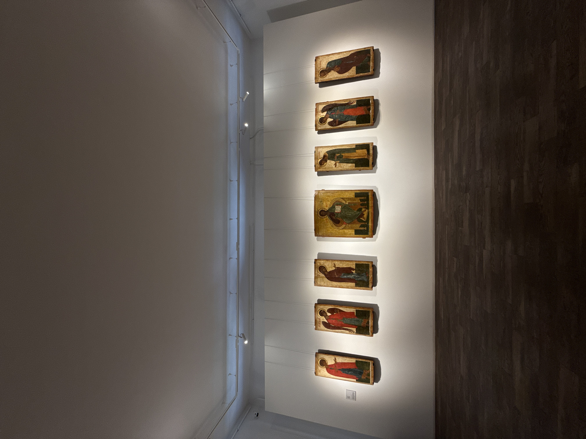 The image captures a scene within the Nizhny Novgorod State Art Museum in Russia. Seven religious icons are vertically aligned on a white wall. The icons are rectangular and depict figures in traditional religious attire. The icons are illuminated by spotlights attached to a track lighting system on the upper left of the wall. To the left of the icons is a large expanse of off-white wall. To the right, a dark brown wall runs vertically, with the appearance of dark wood planks. A small sign is visible below the bottom-most icon. There is no one is present in the frame.