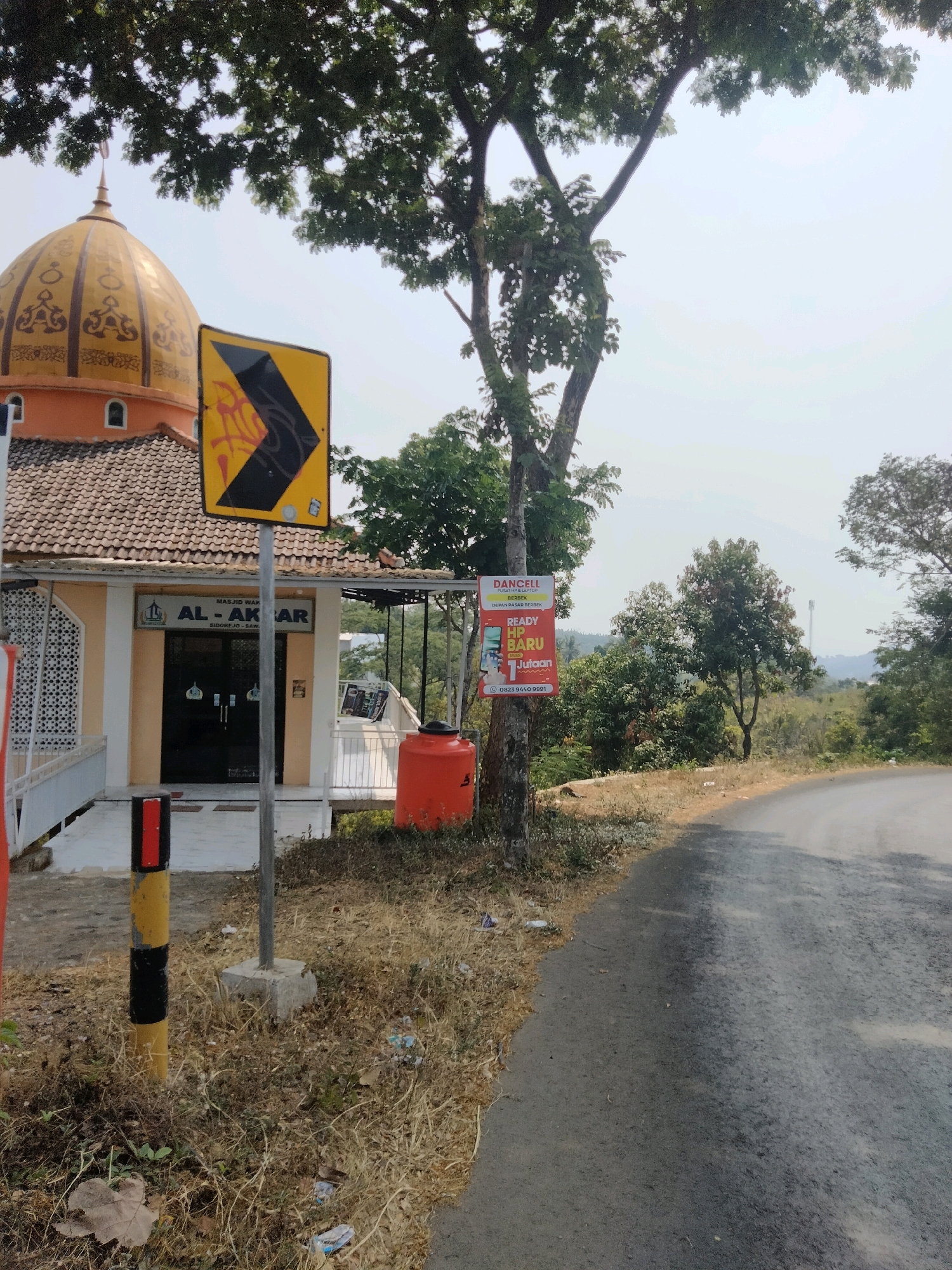 In Sidorejo, Indonesia, a yellow and black directional road sign pointing to the right is posted near a mosque. The mosque, named "AL-AKAR" with the location "SIDO-REJO - SAWAH", has an orange exterior and a golden dome. Adjacent to the mosque is an orange water tank. A tree with green foliage stands behind the sign and the mosque. Further in the background, a road curves to the right, bordered by dry grass and vegetation. The sky is overcast.
