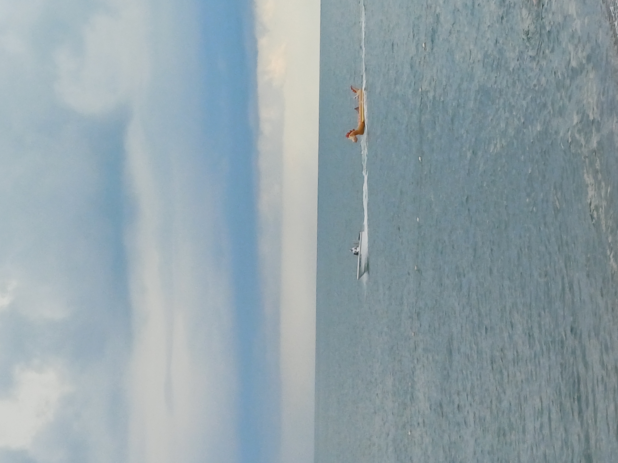 The image captures a wide shot of the Black Sea with a slightly overcast sky over Sochi, Russia. In the foreground, the choppy, blue-grey water fills most of the frame. A small white motorboat, with a person visible at the helm, is seen moving from left to right, creating a white wake. Towed behind the motorboat is a large, inflatable yellow unicorn float with two people riding on it, also leaving a wake. The overall scene depicts a leisurely activity on the water under a cloudy sky.