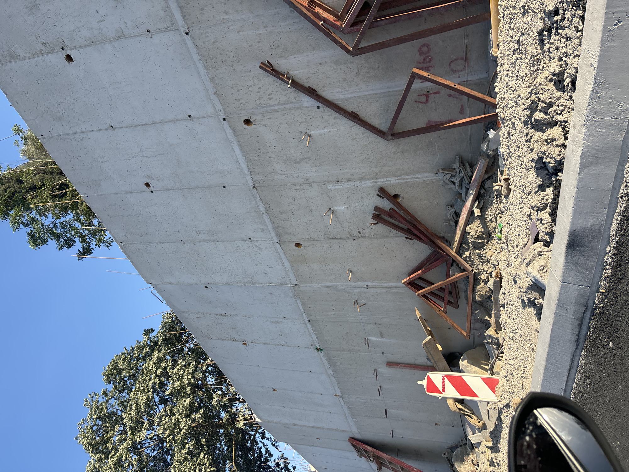 The image captures an outdoor construction scene, primarily featuring the underside of a large, unfinished concrete structure, likely a bridge or overpass. The gray concrete slab extends diagonally across the frame, displaying formwork lines, numerous small holes, and several embedded or attached rusty metal ties and rebar.
To the right of the concrete structure, the ground is covered in loose, light gray gravel and construction debris, including discarded sections of rusty metal frames and other refuse. A white and red striped barrier or sign is visible in the lower right, partially obscured by a car's side mirror, indicating the photograph was taken from a vehicle. The word "BEMAX" is printed on this barrier. Faintly visible on one of the rusty metal frames near the top right are the numbers "4160."
In the background, a clear, bright blue sky is visible, along with the green canopy of several trees, suggesting a natural or semi-urban setting. The bright illumination indicates it is daytime, likely a sunny day. The overall scene conveys an active construction environment, with exposed concrete and materials characteristic of ongoing infrastructure development.