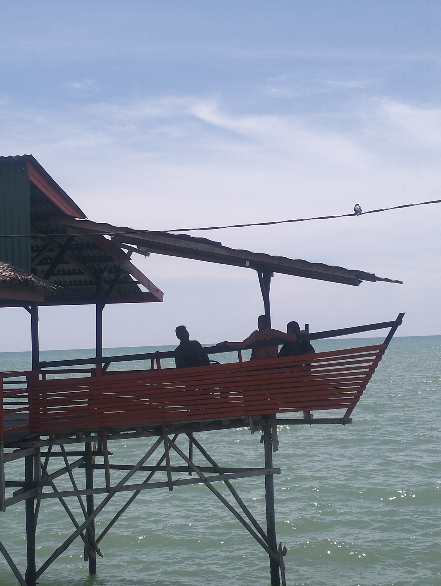The image depicts a stilt house located in Lancok, Indonesia, situated over the sea. The structure features a red wooden deck with horizontal slats and a covered roof with green corrugated metal and traditional thatched roofing. Three individuals are visible on the deck, silhouetted against the bright sky, appearing to be seated or standing. The sea is calm with gentle ripples reflecting sunlight. In the upper right portion of the image, a bird is perched on a thin wire stretched across the sky. The sky is predominantly blue with scattered, wispy white clouds. The overall scene is characteristic of a seaside dwelling or rest stop in the coastal region.