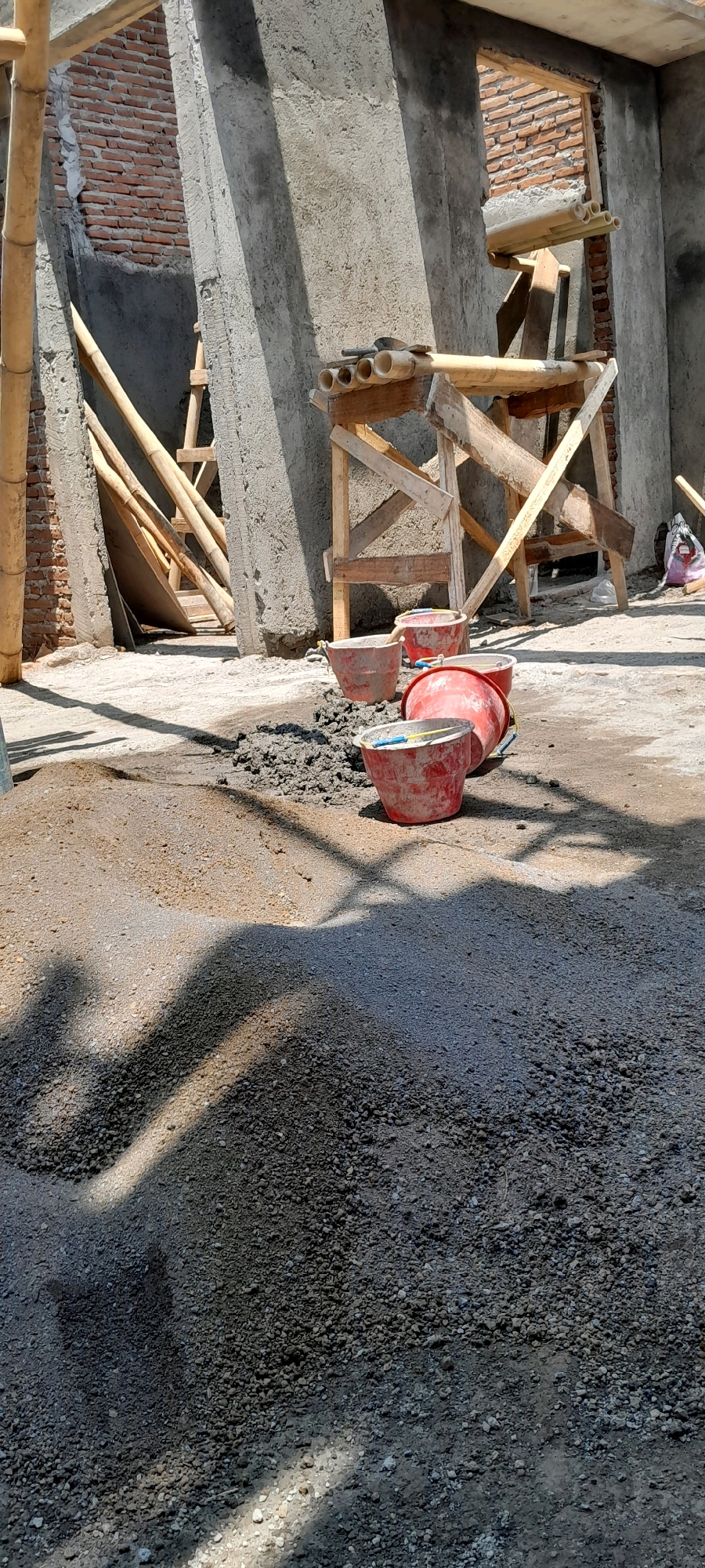The image depicts a construction site in Karangduren, Indonesia, with building materials and unfinished structures. In the foreground, a pile of sand and gravel is visible, with several red buckets scattered around it. Some of the buckets contain cement, while others appear empty. In the background, brick walls, concrete pillars, and bamboo scaffolding are present, indicating ongoing construction work. The scene is illuminated by sunlight, casting shadows on the ground. The overall impression is one of a typical construction environment, with elements of building materials, tools, and architectural elements.