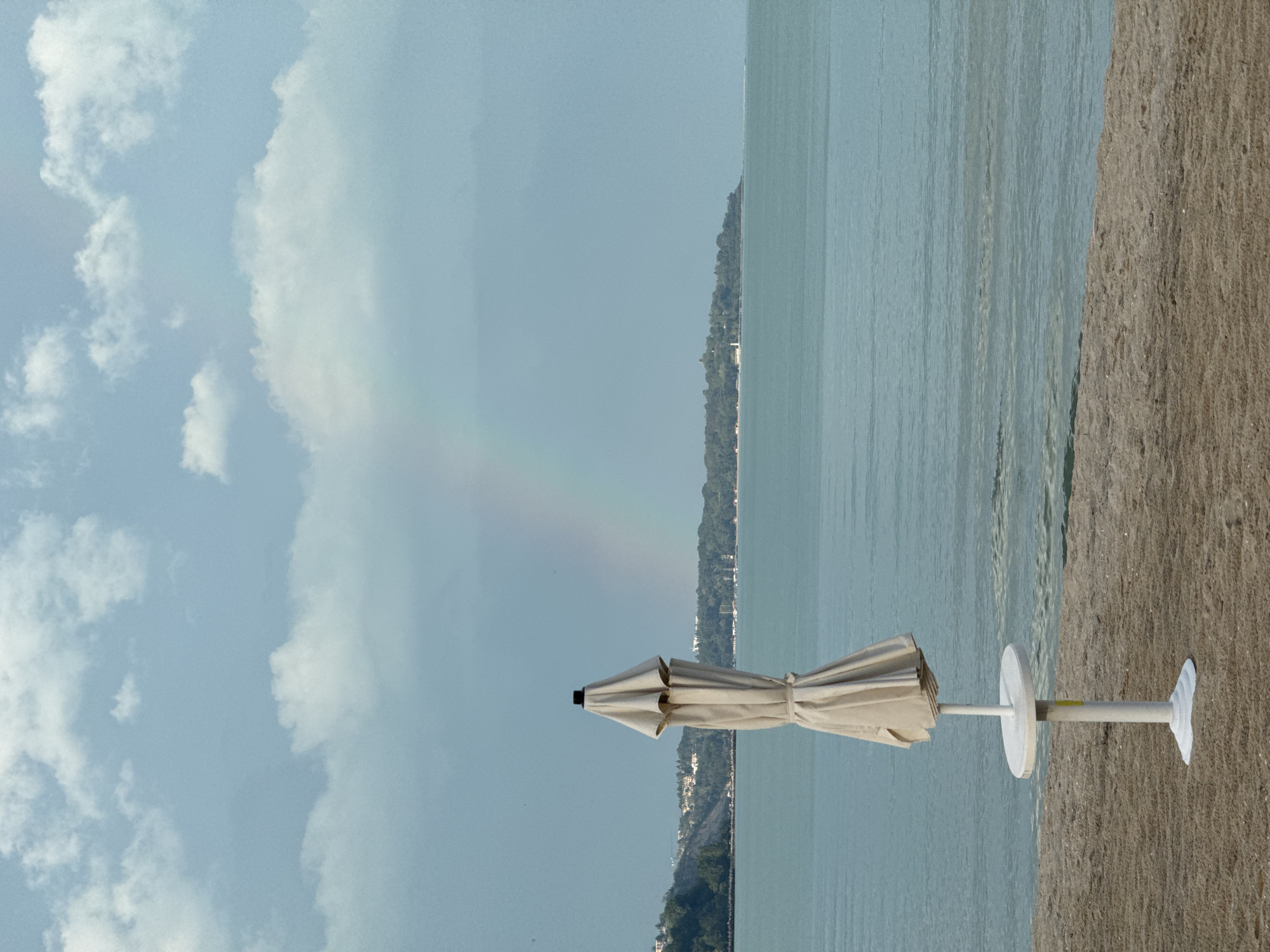 The scene shows a beach view with an unopened beige beach umbrella in the foreground, its base stuck in the sand. Behind the umbrella, there's a body of water, possibly the Black Sea. Across the water, a landmass with green trees and buildings lines the horizon. In the sky, there's a visible rainbow arc under some clouds. The image is taken from a location on the beach in Varna, Bulgaria.