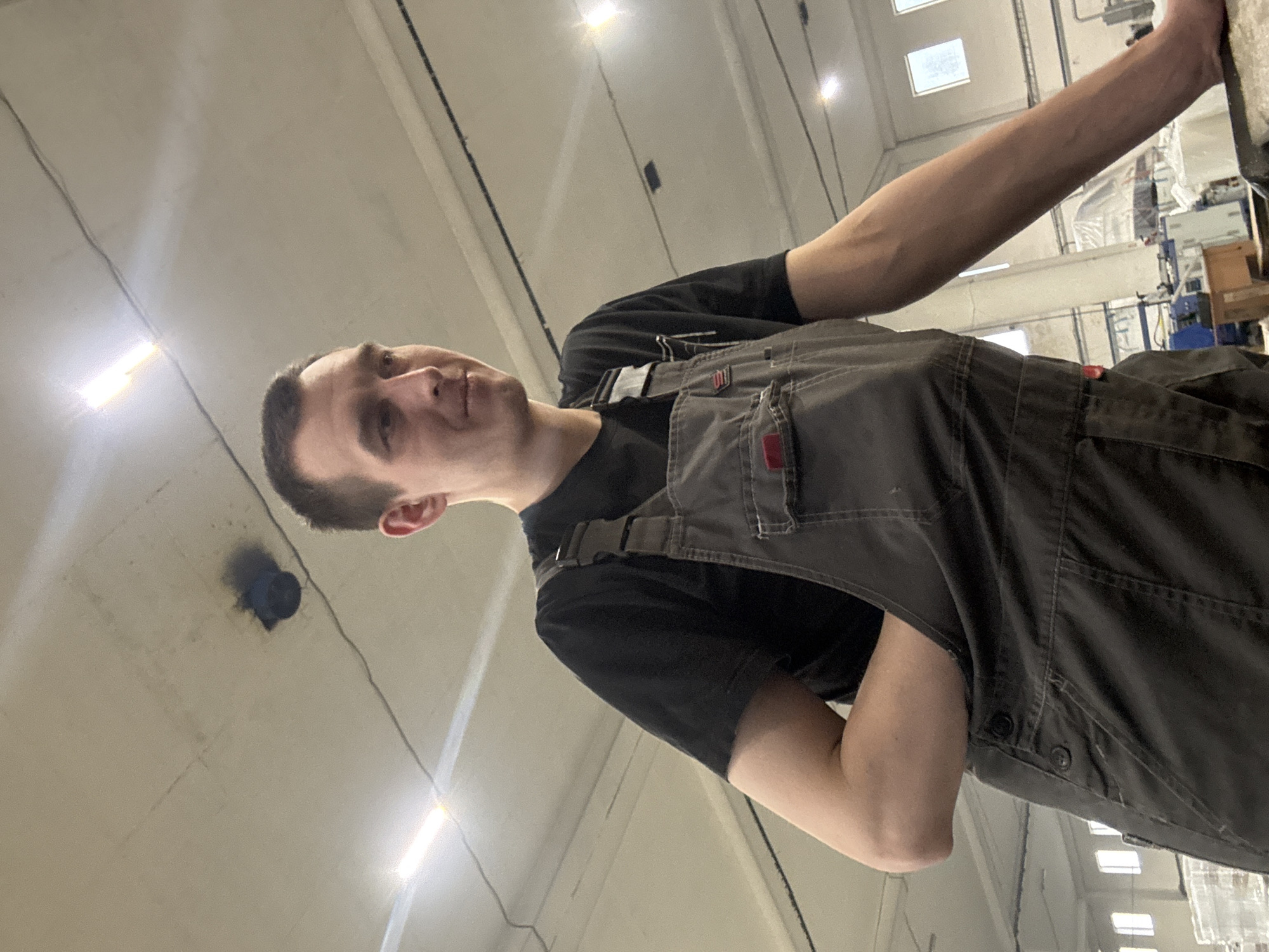 The image shows a male individual in what appears to be an industrial or workshop setting located in Dikaya Gar', Russia. The man, an adult with short dark hair, is viewed from a low angle, facing slightly to his right with his gaze directed towards his right. He is wearing a black short-sleeved t-shirt beneath dark grey work overalls, which have visible buckles on the left shoulder strap and buttons on the left side. A pen is clipped to the chest pocket of his overalls. His left arm is bent, with his hand inserted into the front pocket of his overalls. His right arm is extended and rests on an unseen surface to the right of the frame. The background features a high, light-colored concrete ceiling with visible seams, exposed electrical wiring, and bright fluorescent strip lights providing artificial illumination. A dark blue, cylindrical object, possibly a vent or pipe opening, is visible on the ceiling with dark smudges surrounding it. Through windows in the distant background, natural light is apparent, and various industrial equipment or stacked white, translucent objects are partially visible below the windows. A blue object and a wooden box are also discernible as part of the background machinery or equipment.