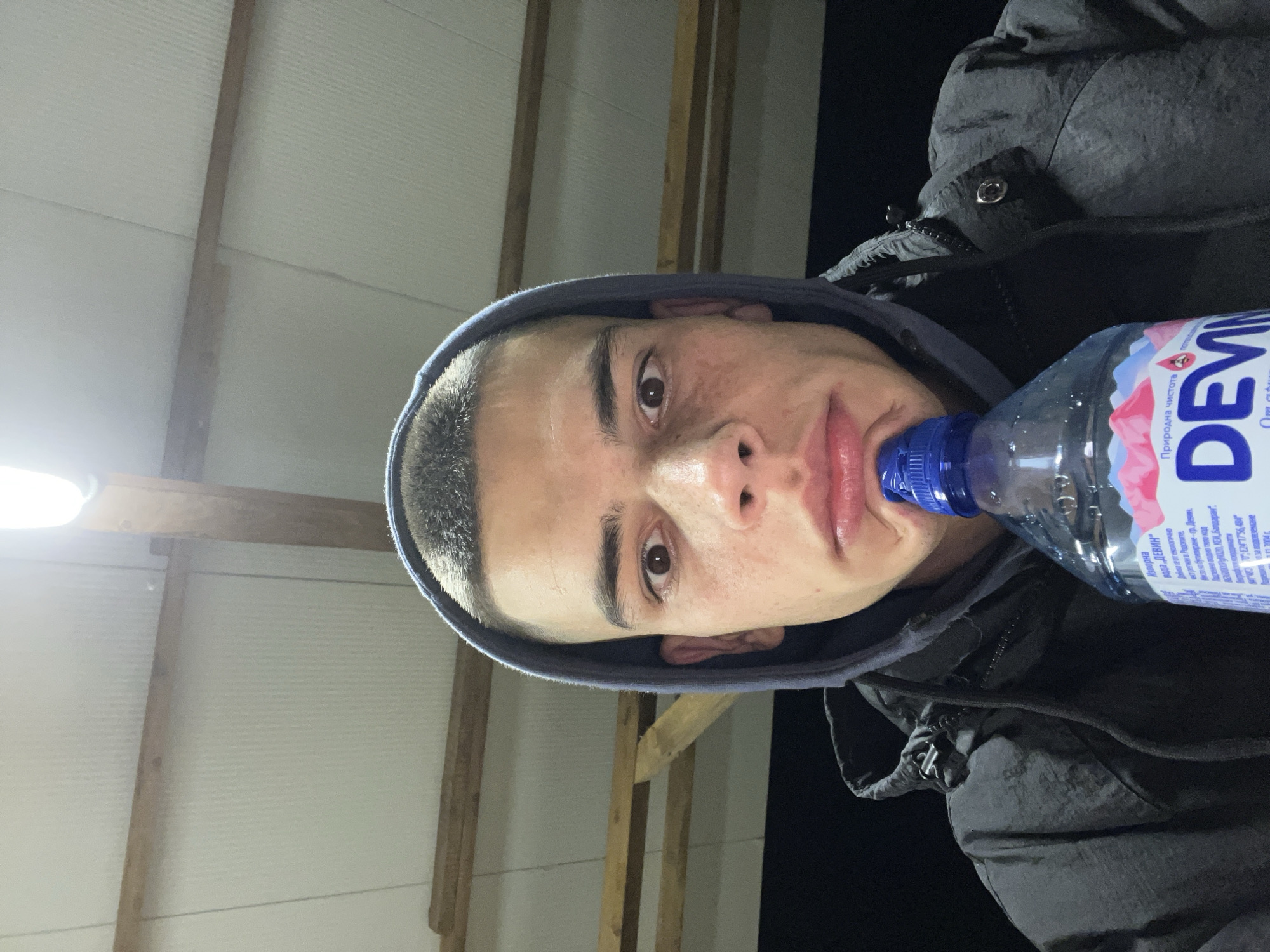 A young man with short, dark hair, wearing a grey hooded jacket, is holding a plastic bottle of "Devin" brand water to his lips. The label on the bottle is in Cyrillic. The background is partially visible, showing a corrugated white wall and what appears to be wooden beams. The lighting is uneven, with a bright light source on the left side of the frame. It cannot be determined whether the image was taken in Karavelovo, Bulgaria from the information provided.