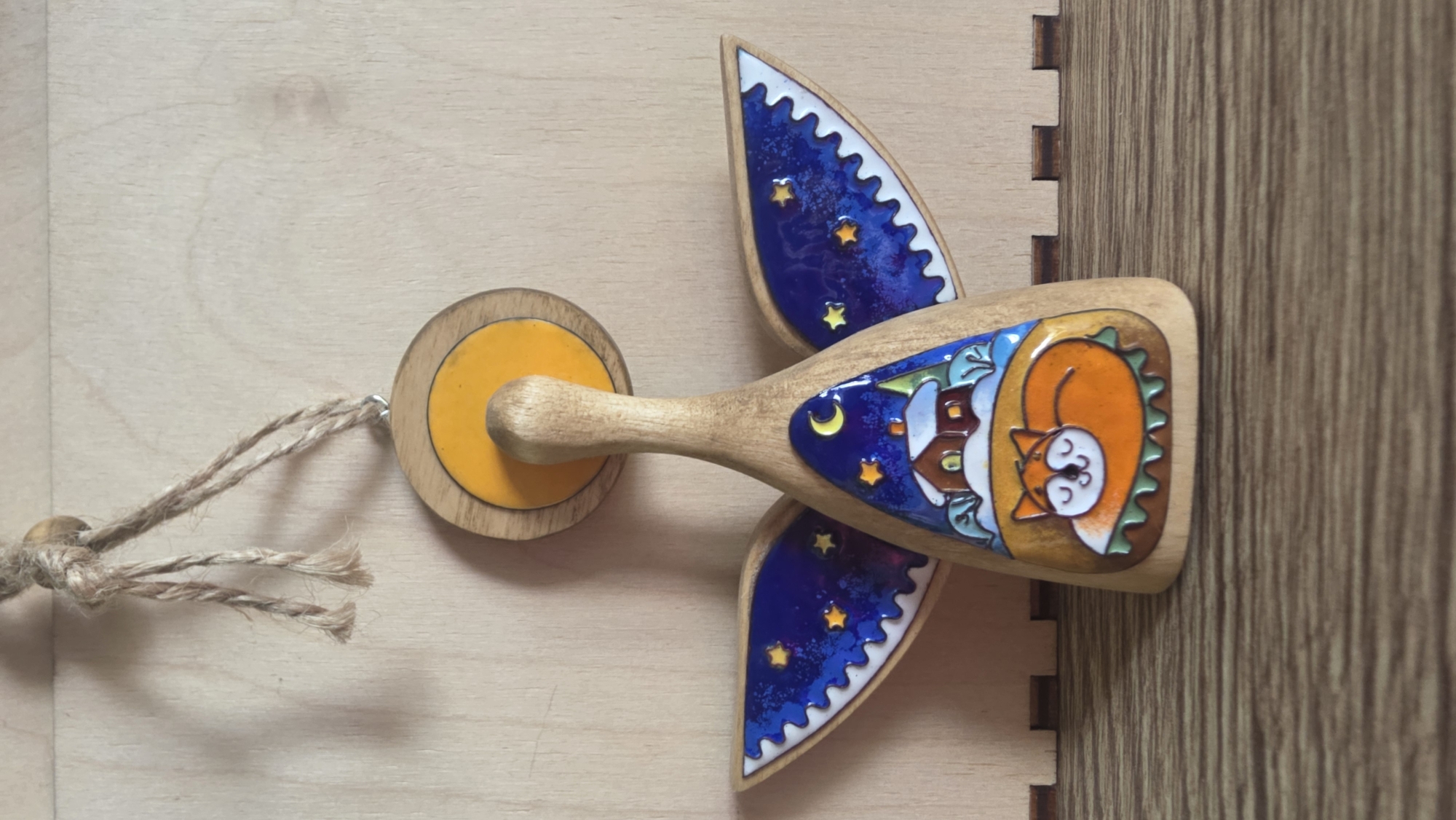 A close-up shot displays a wooden toy, likely a decoration, featuring an orange cat curled up next to a house under a starry sky, with blue wings decorated with stars. The toy is resting on a wooden surface. The picture appears to be taken indoors. It is not possible to identify a location in Saint Petersburg, Russia based on this image.