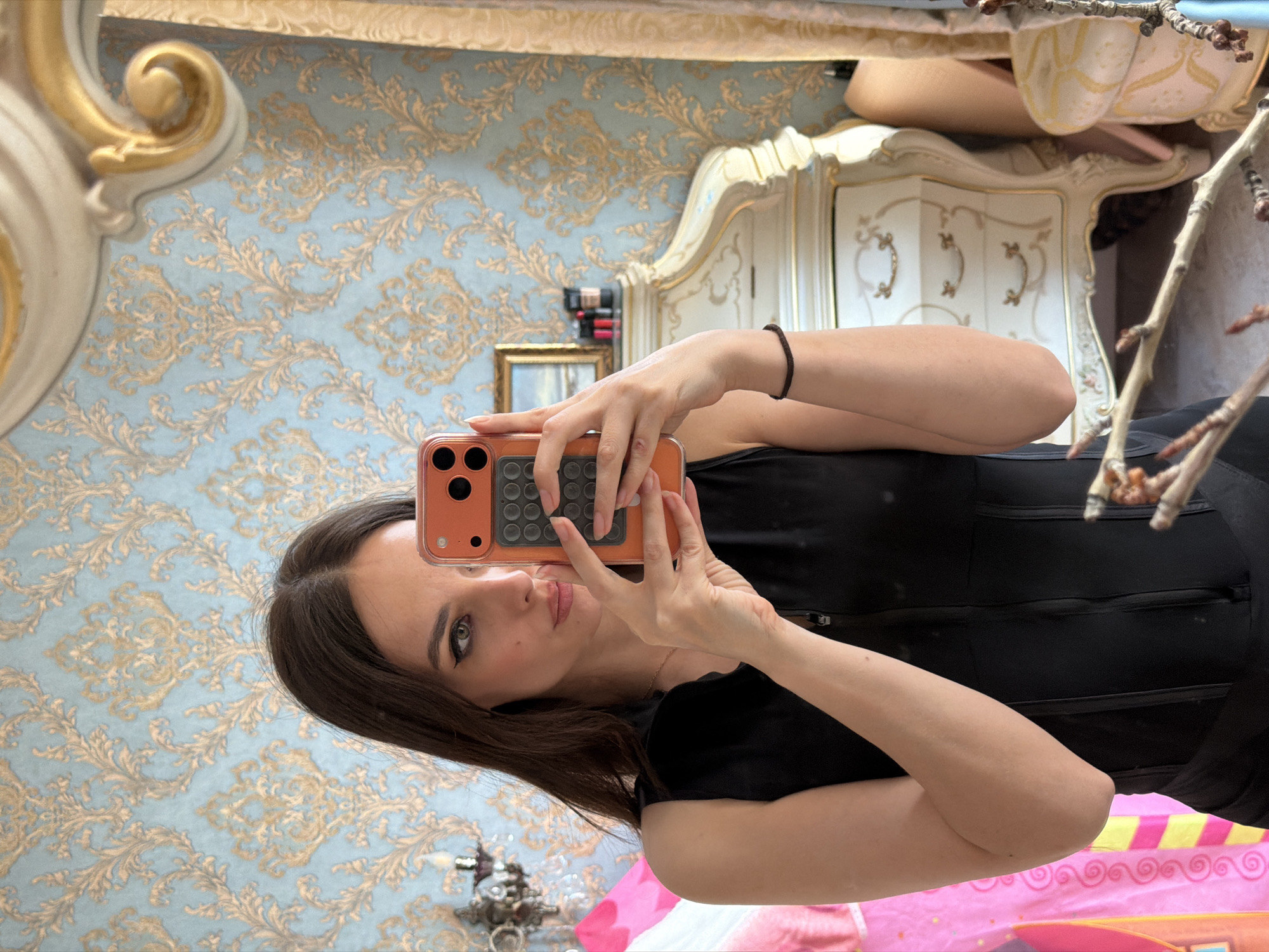A woman takes a mirror selfie in an ornate room. She is wearing a black top and her right arm is bent at the elbow as she holds her phone. The phone has an orange case and is positioned to capture her reflection. Her dark hair is down, and she is looking at the phone with a neutral expression.
The room has ornate, blue and gold patterned wallpaper. A large, gilded mirror frame with intricate carvings is visible to the left of the woman. To her right, a white and gold cabinet with decorative carvings is partially visible. The overall impression is of a luxurious and classic interior. There are no visible cues about the time of day or weather. No text is visible in the image.