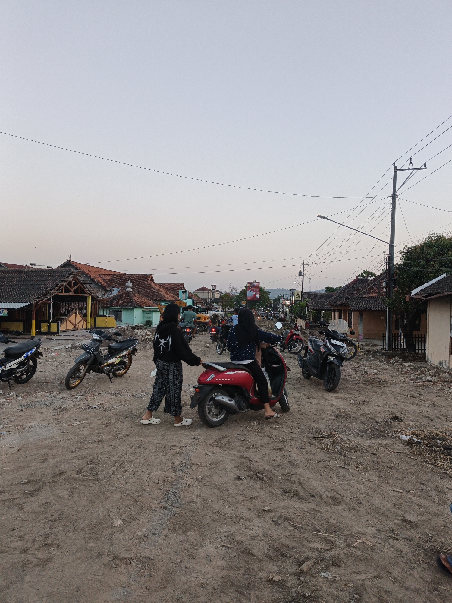 The image depicts a dusty, unpaved street scene in what appears to be a rural or semi-urban area of Planggu, Indonesia, at dusk. Several motorcycles are parked and in motion. Two young women are the central figures, one standing next to a red scooter, facing another woman seated on the same scooter. The standing woman is wearing a black hoodie with a white graphic and patterned pants, while the seated woman has dark hair and is wearing dark clothing. The environment is characterized by rough, dirt ground with scattered debris and construction materials, suggesting ongoing road work or development. Buildings with tiled roofs line the street, some appearing weathered or under renovation. Utility poles and wires crisscross the sky, indicative of a developed but potentially older infrastructure. The overall atmosphere is quiet, with the soft light of late day casting long shadows.