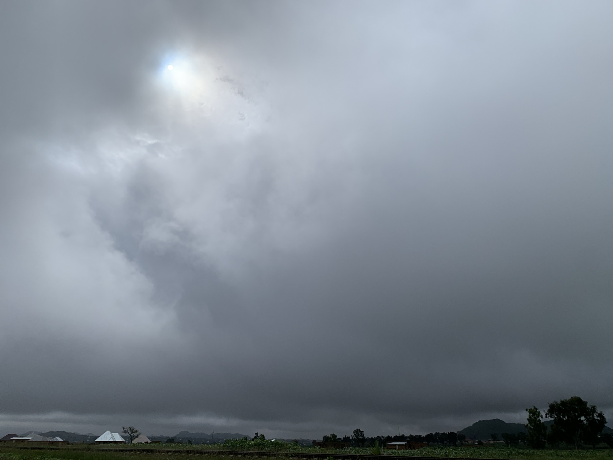 The image depicts a cloudy sky with a bright sun peeking through in the upper left corner. In the foreground, there is a flat landscape with a few houses and trees visible. A railway line runs horizontally across the middle of the image. The overall impression is of an overcast day with a hint of sunlight. The location is identified as Kuru Karama, Nigeria.
