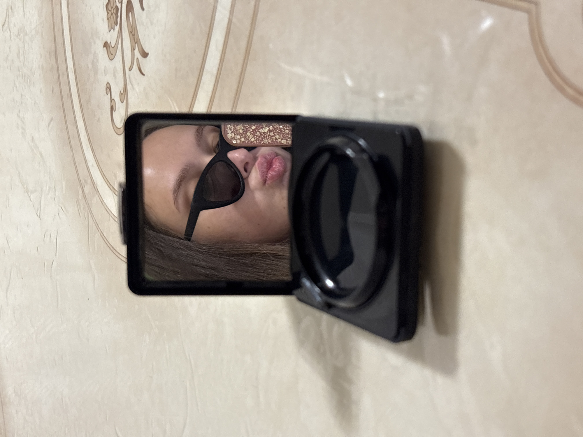 A photograph depicts a woman reflected in the mirror of a small, black compact makeup case attached to a wall. The woman is wearing sunglasses and has pursed lips. The wall appears to be beige with a faint, patterned design. No further location information is discernible from the image.