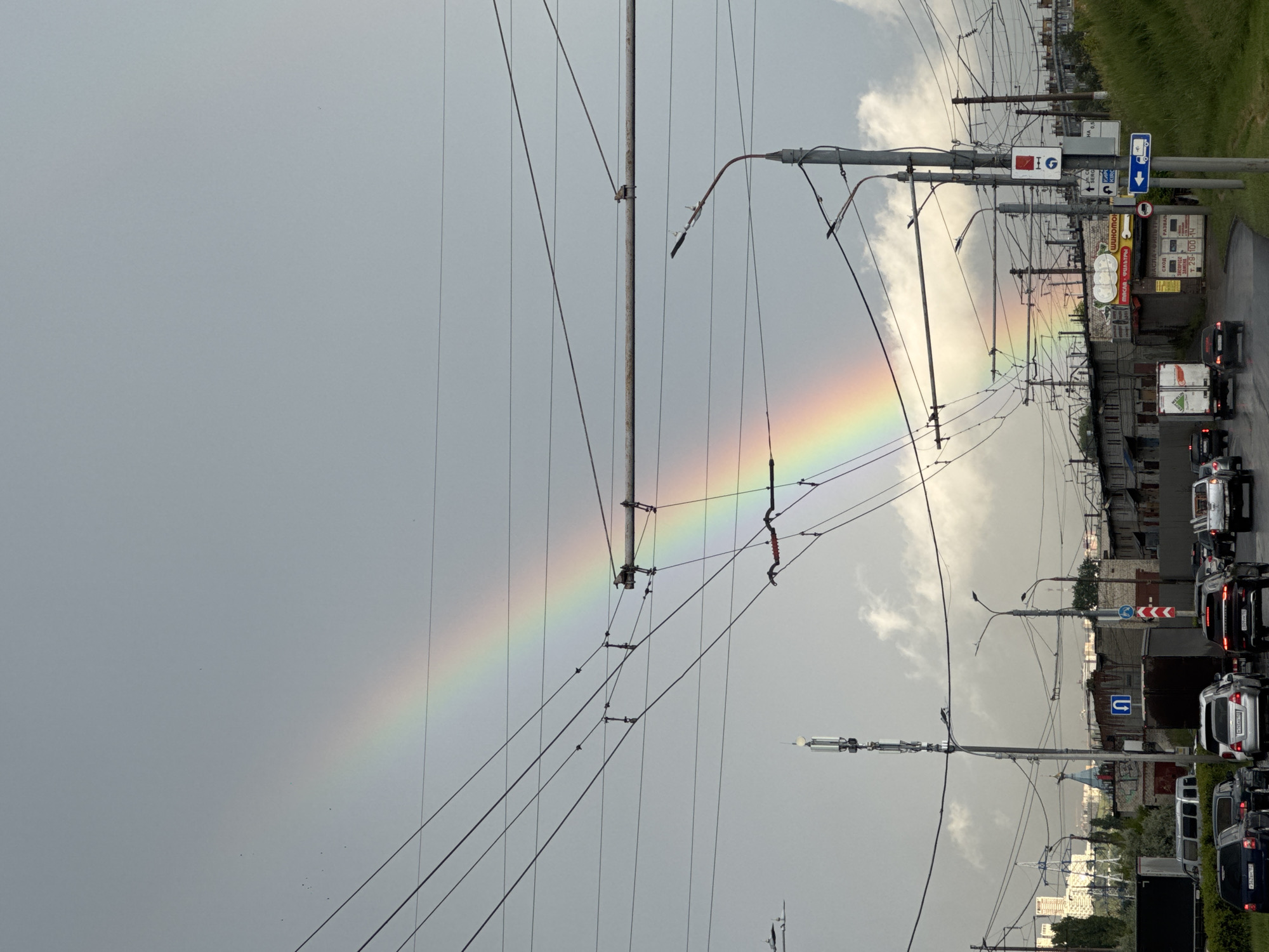 The media file depicts a city street scene, rotated 90 degrees counter-clockwise, with a prominent rainbow arching across a cloudy sky. The foreground is dominated by a dense network of overhead wires, likely for trolleybuses or trams, supported by tall poles. These wires create a web-like pattern against the pale, overcast sky.
To the right of the image, a street is visible with several vehicles, including cars and trucks, suggesting traffic. Buildings and some greenery line the street, with architectural styles indicating an urban environment. Signage is visible along the street, some of which are in Cyrillic script. One sign has a blue arrow indicating a U-turn. Another sign appears to be a speed limit sign, possibly indicating 40. A sign with red and white markings and an icon of a person walking is also visible. Some shops or businesses with Cyrillic text are present, including one with "МЯСО - ФИЛИАЛ" and another that says "ШИНОМОНТАЖ".
The presence of the rainbow suggests recent or ongoing rain, though the sky is mostly cloudy. The overall mood is somewhat somber due to the overcast conditions, but the rainbow adds a touch of beauty and hope to the scene. The time of day appears to be late afternoon or early evening, based on the lighting and cloud cover. The location is identified as Nizhny Novgorod, Russia. There are no visible people in the image. The primary subjects are the overhead wires, the rainbow, and the urban street with traffic.