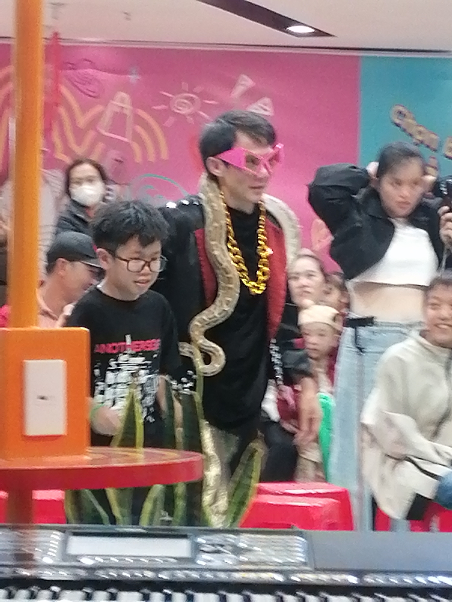A man with a snake around his neck, wearing pink glasses and a gold chain, stands among a crowd of people. A boy in glasses stands in the foreground. The scene appears to be in a public space, possibly a mall or entertainment venue in Da Lat, Vietnam, based on the background design.